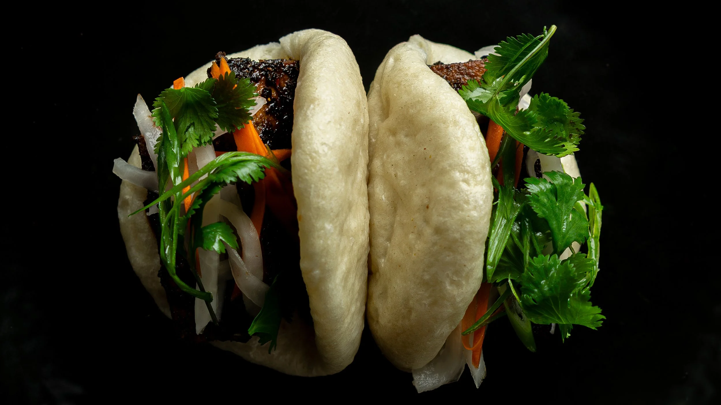 Close-up of two white tacos filled with grilled meat, vegetables, and fresh cilantro against a black background.