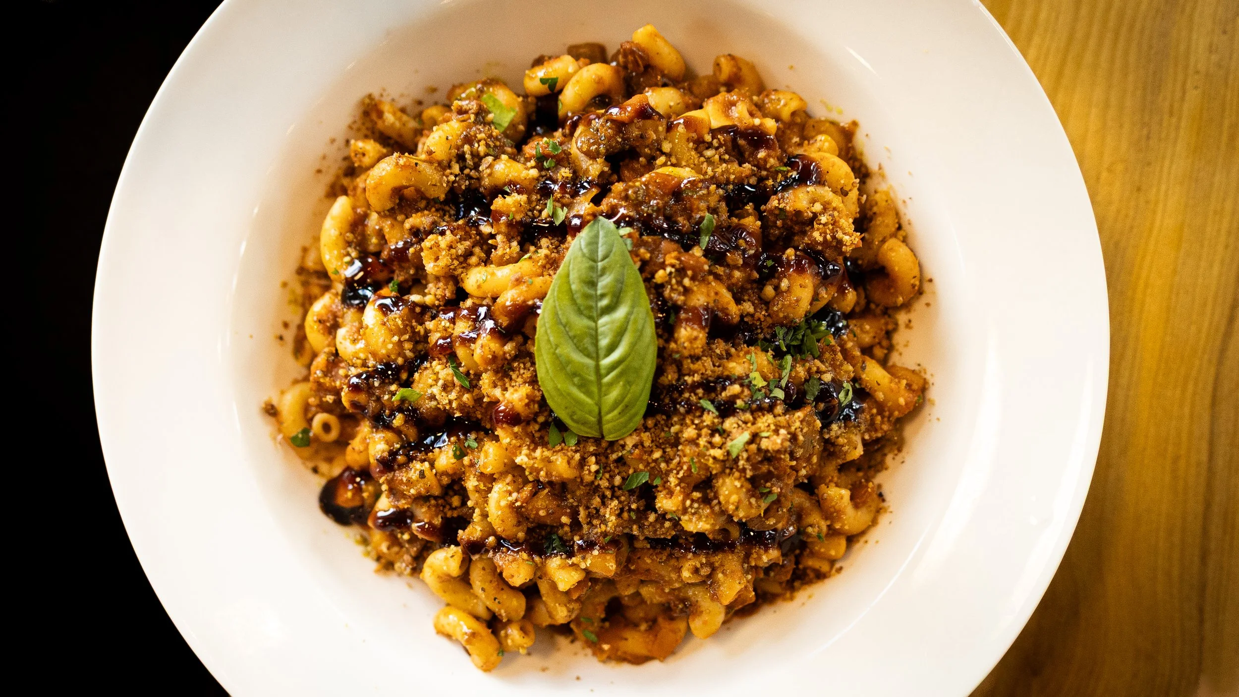Close-up of a bowl of pasta topped with crushed nuts, herbs, and a drizzle of balsamic glaze, garnished with a fresh basil leaf.