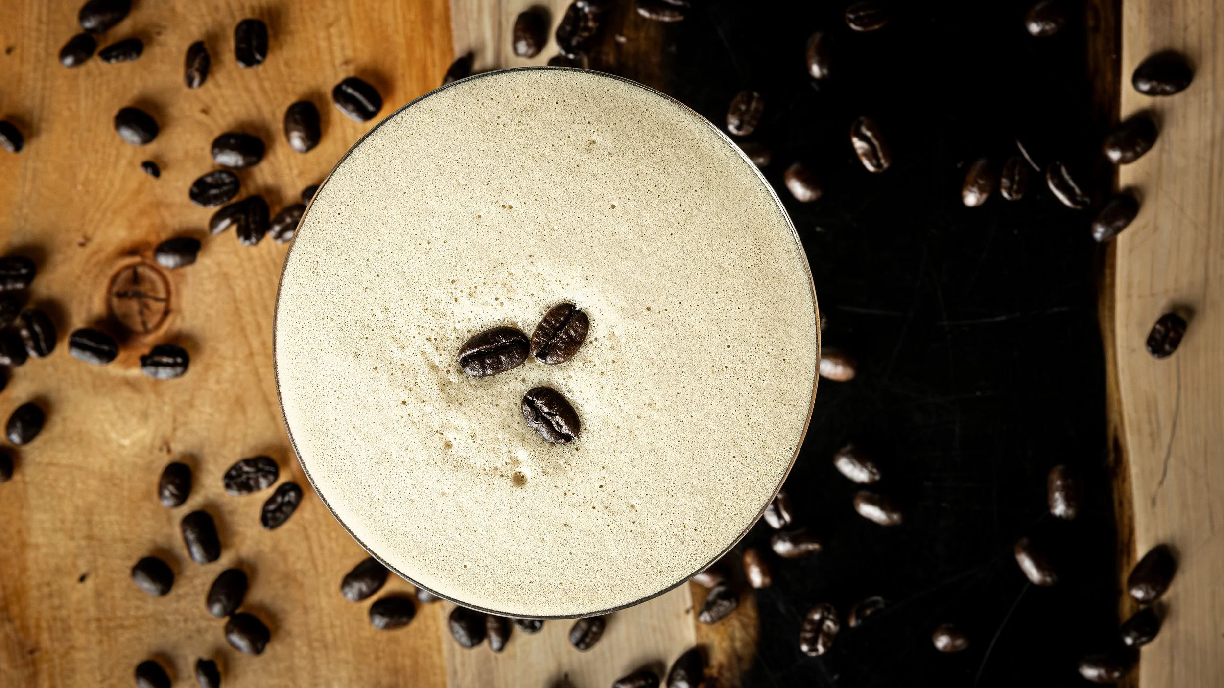 Close-up of a frothy coffee drink with three coffee beans on top, surrounded by scattered coffee beans on a wooden surface.