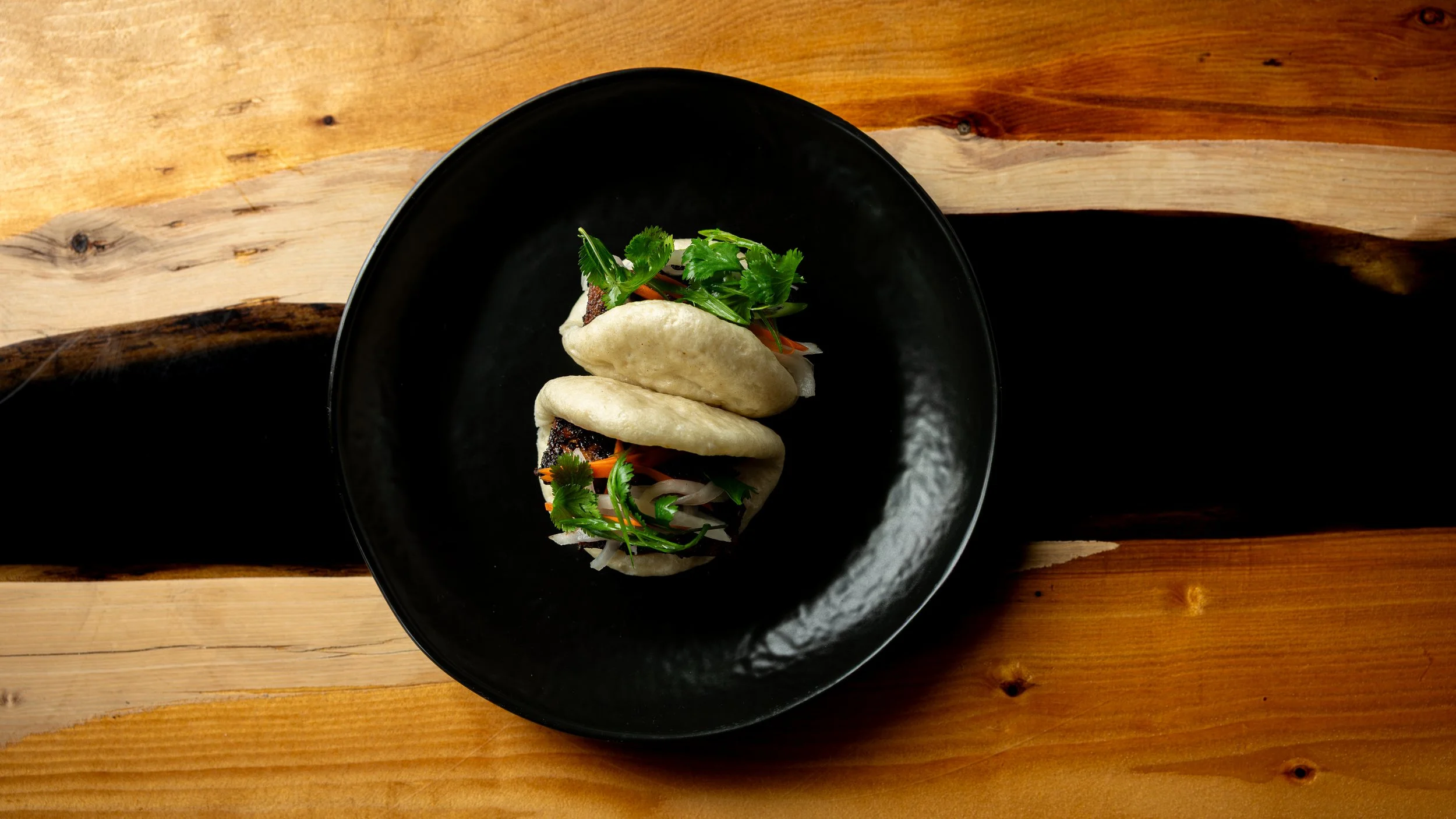 A black bowl containing two steamed bao buns filled with grilled meat, fresh cilantro, and sliced vegetables, placed on a wooden table.