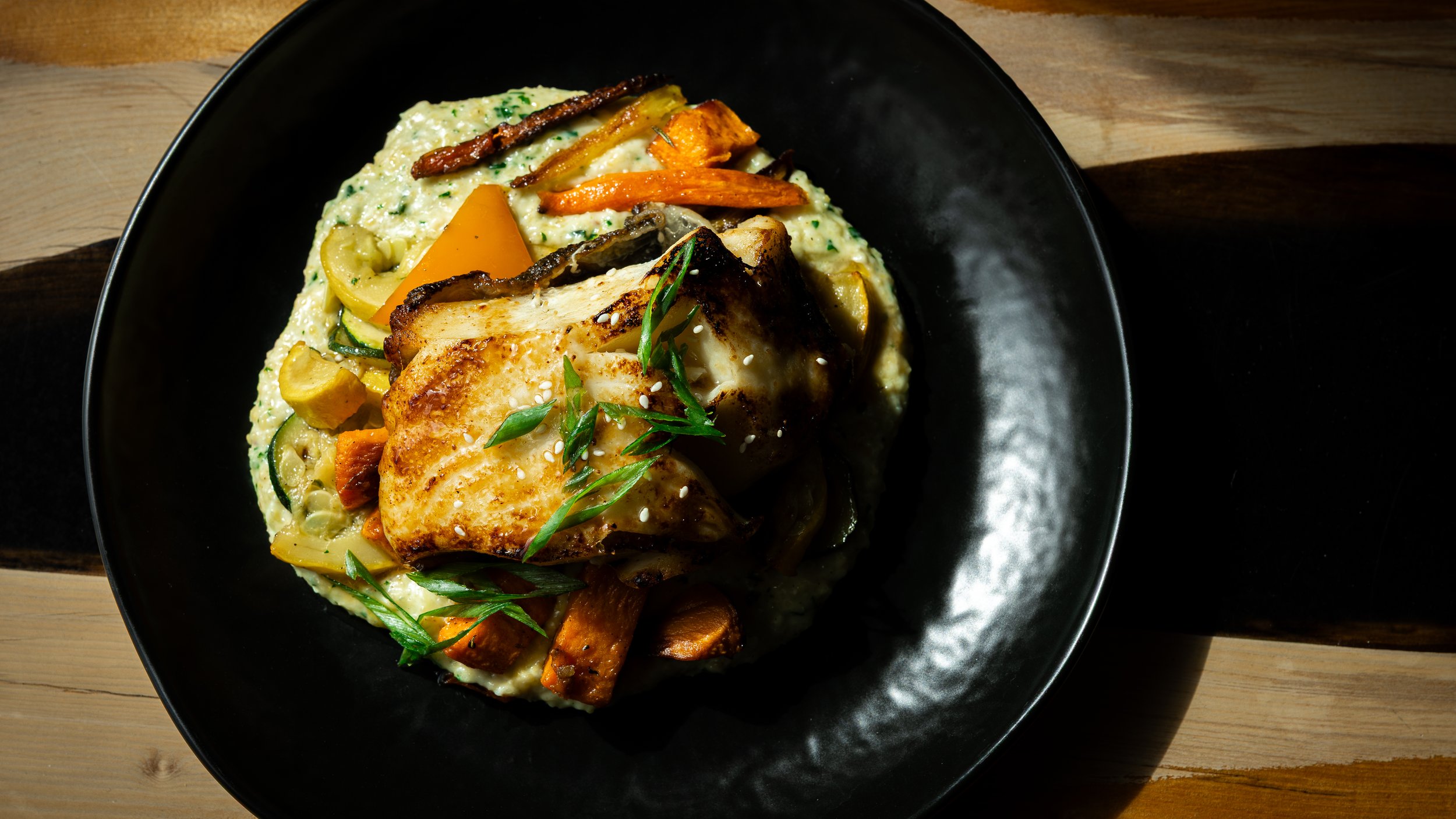 A black bowl with cooked fish fillet, surrounded by sautéed vegetables and garnished with green herbs.