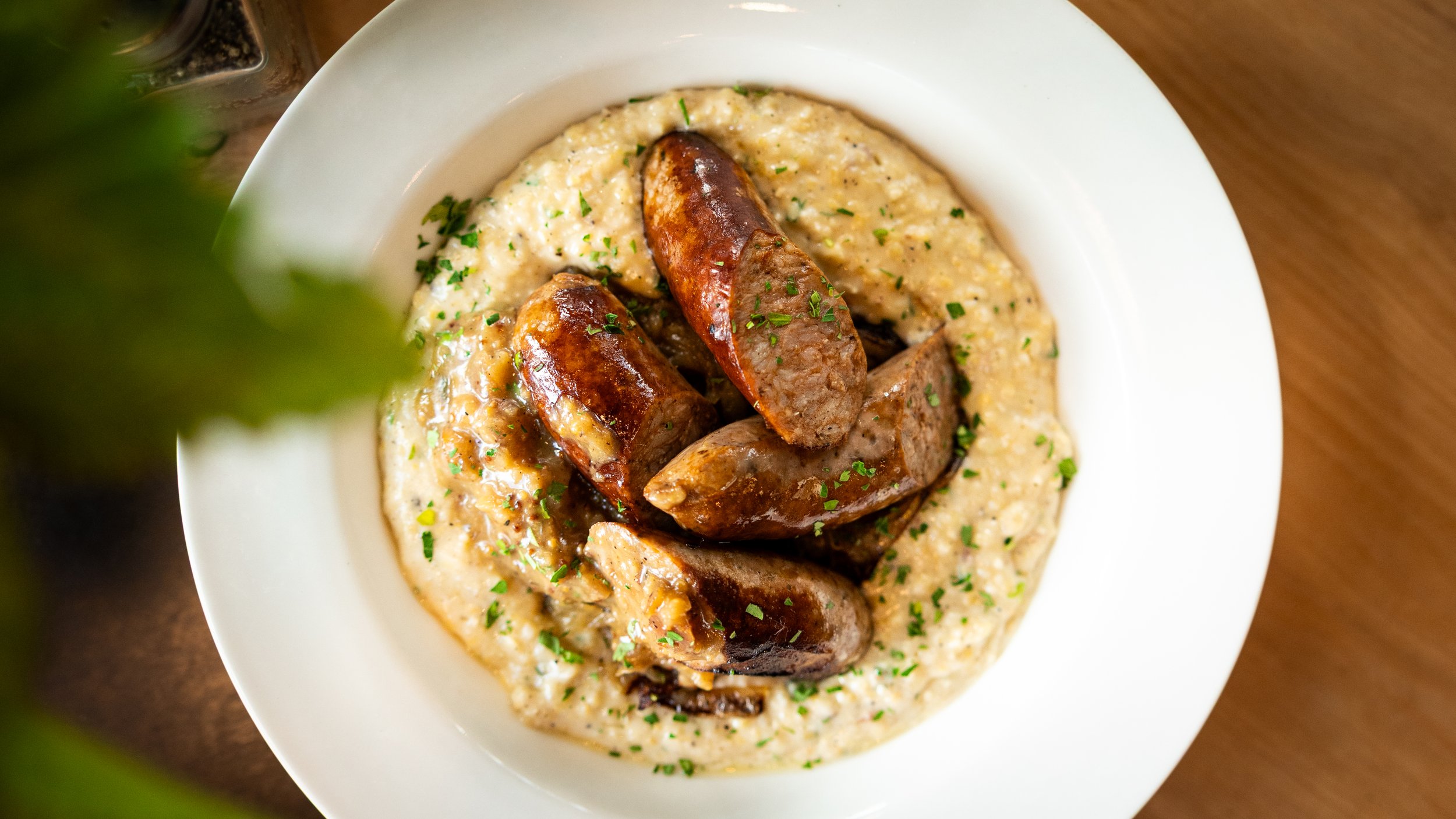 Plate of creamy mashed potatoes garnished with chopped parsley, topped with several chunks of cooked sausage or meat, drizzled with sauce.