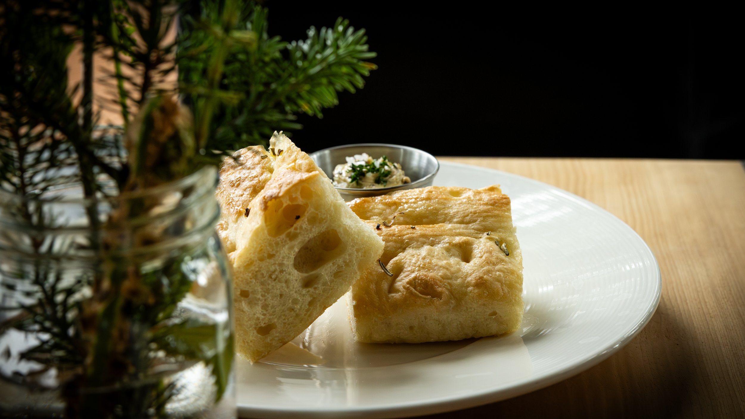 Close-up of a white plate with pieces of bread or focaccia on a wooden table, with a small bowl of herb butter or spread in the background, and a blurred plant in a jar in the foreground.