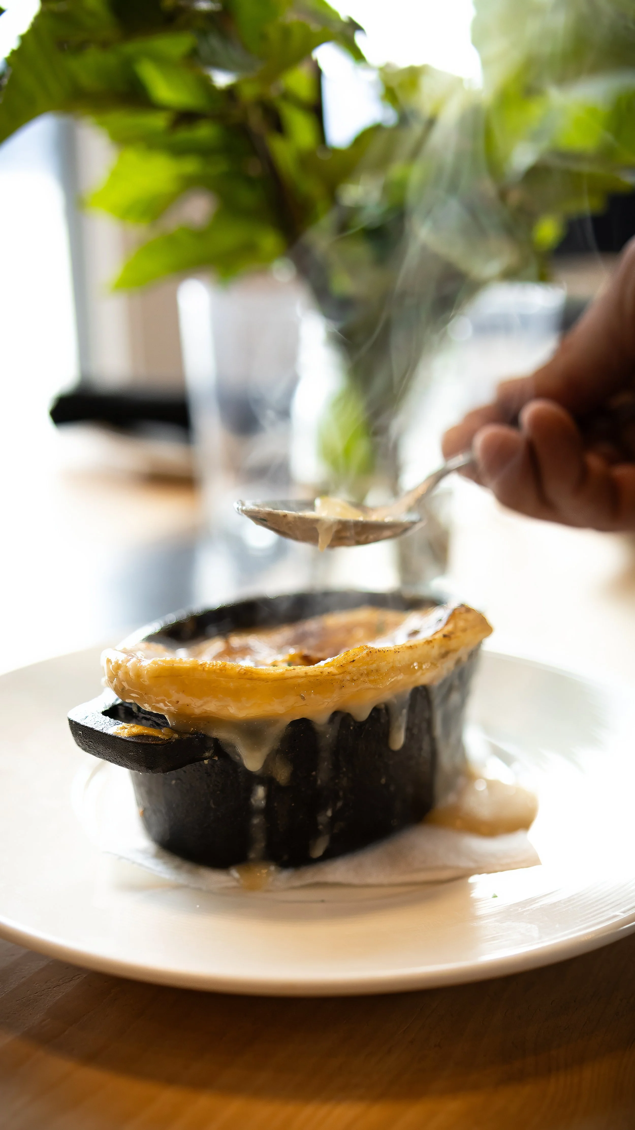 A dish with a baked egg in a black dish, topped with melted cheese, and a hand holding a spoon above, in a setting with natural light and green plant in the background.