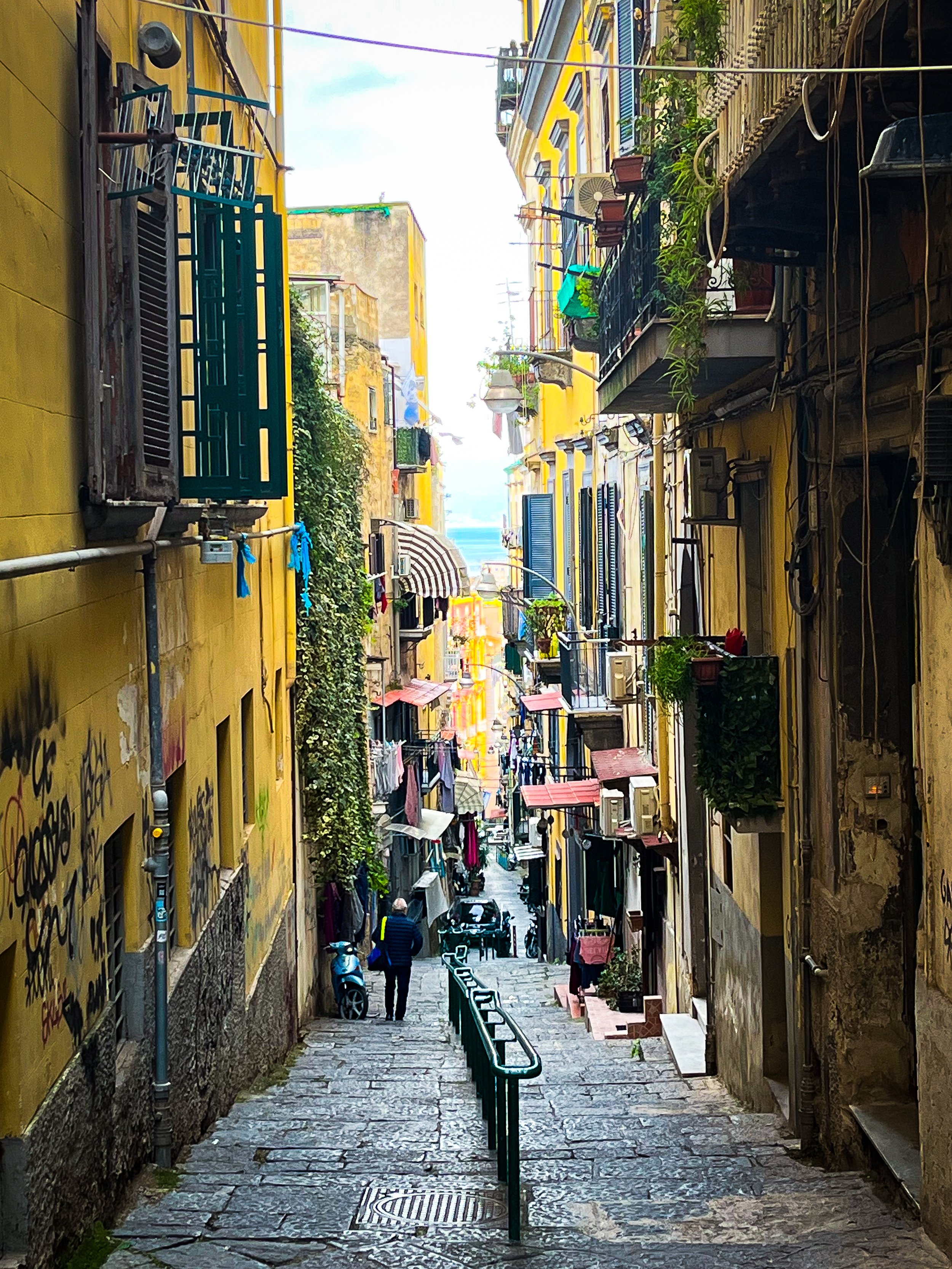Narrow alleyway with graffiti-covered yellow walls on the left and apartment buildings with balconies on the right, leading down to the street with parked scooters and a person walking.