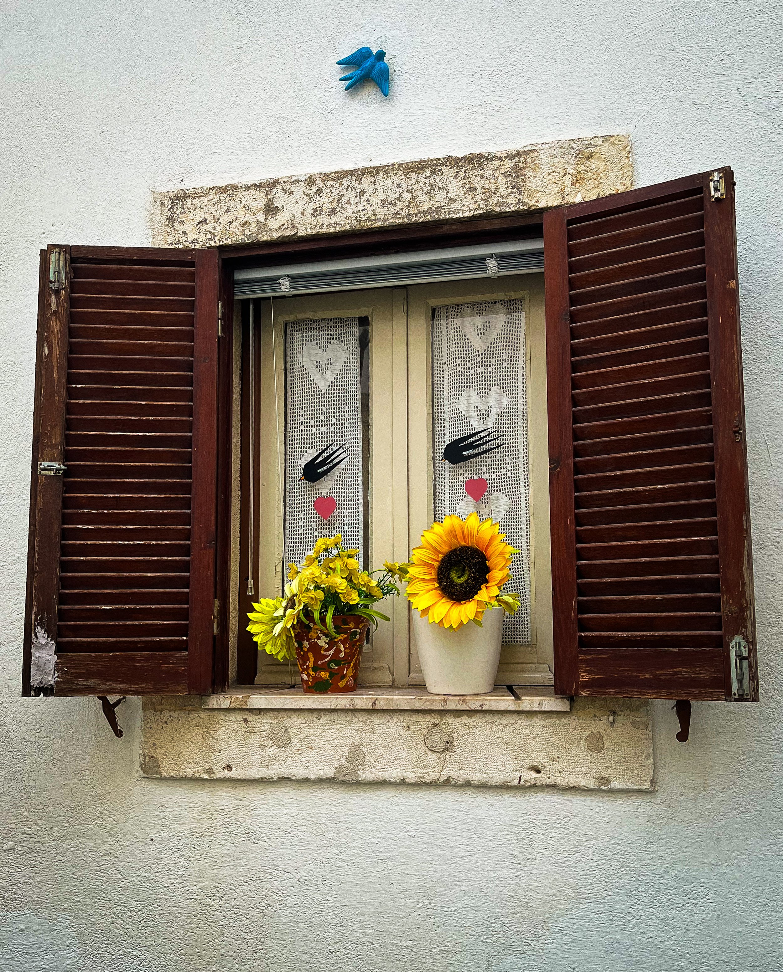 A window with wooden louvered shutters open, set in a white textured wall. On the window sill, there are two potted plants with large yellow sunflowers, and lace curtains with heart and hand motifs. Above the window, a decorative blue bird ornament is mounted on the wall.