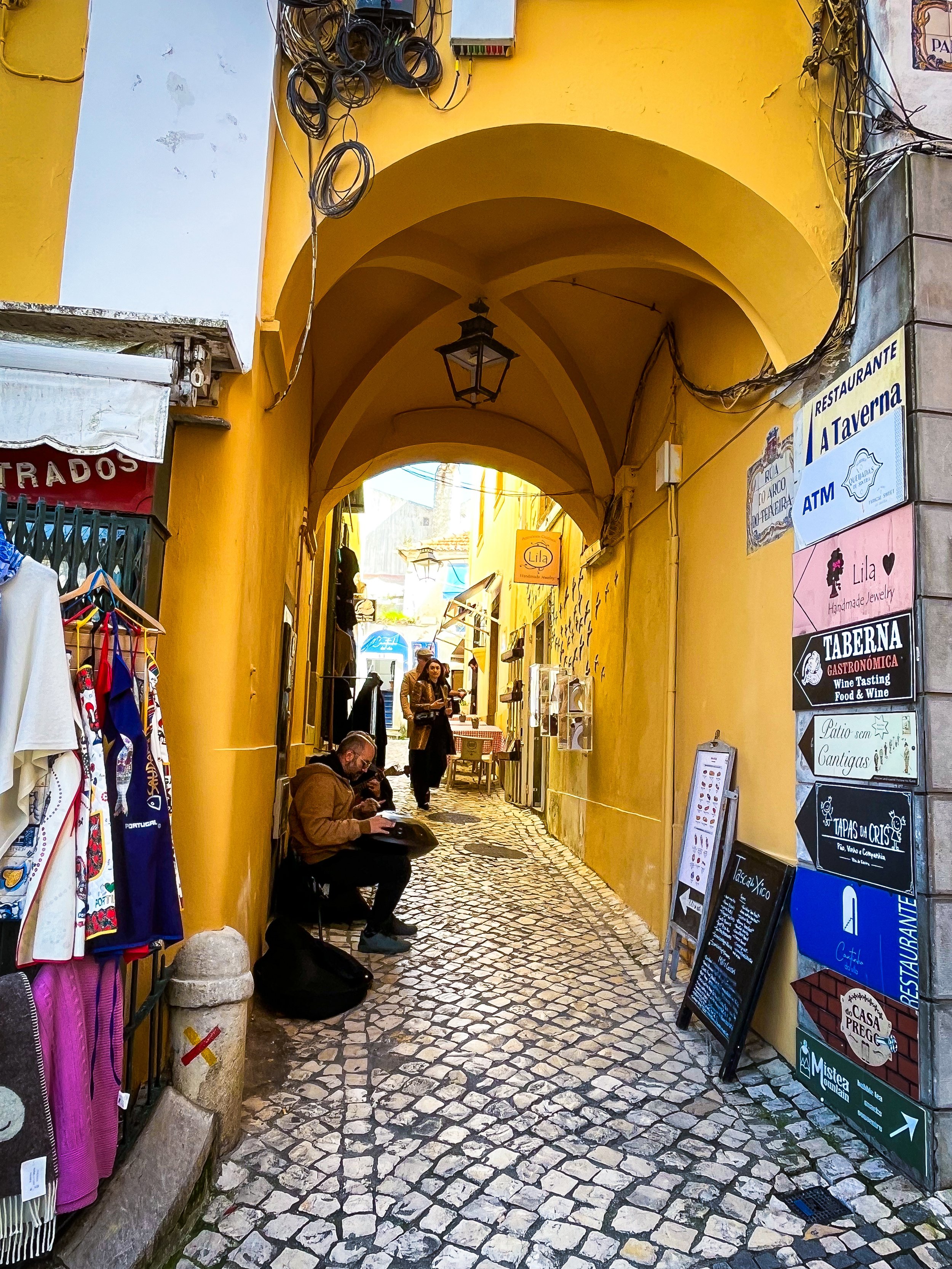 A narrow cobblestone street with yellow walls and arched ceilings. People are sitting and walking, shops and signs are along the street, and a man in a brown jacket is seated on a small stool.