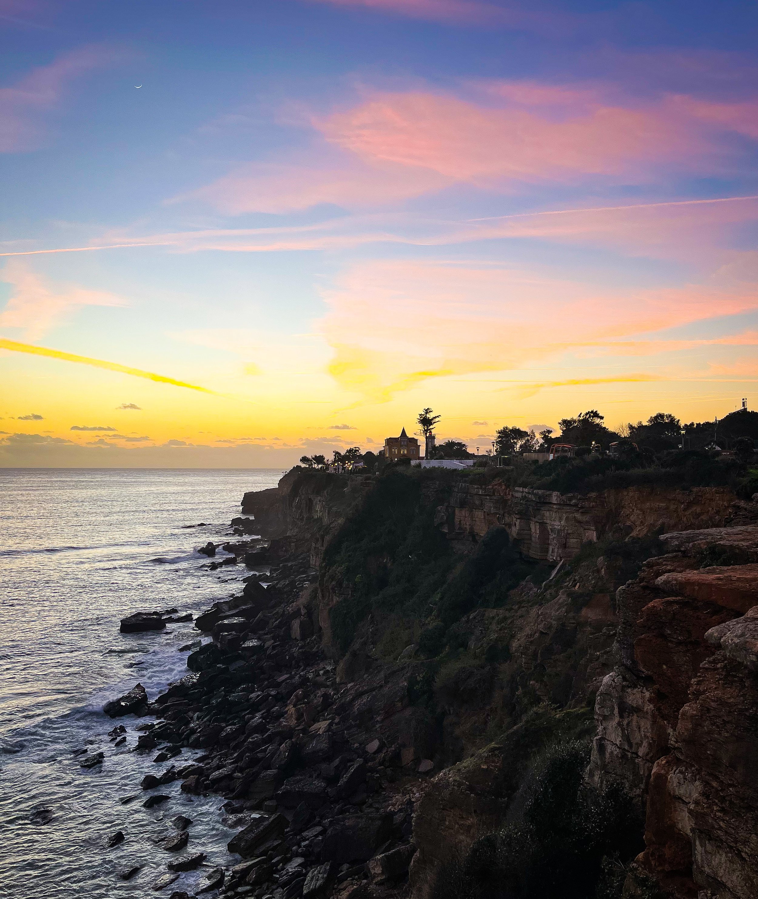 Sunset over the ocean with a rocky shoreline and a silhouette of a house on a cliff, featuring pink and purple clouds in the sky.