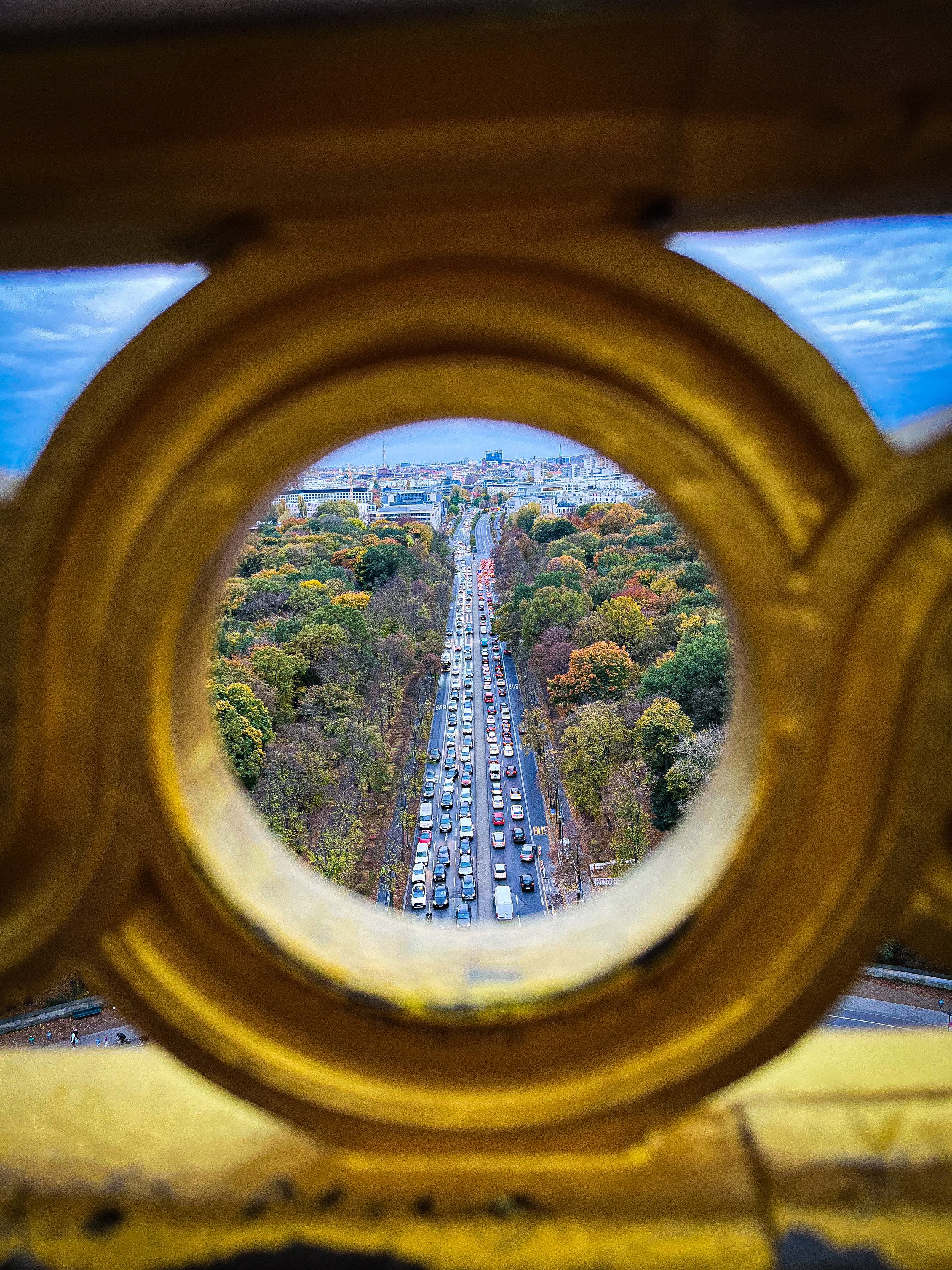 A view through a circular opening in a yellow railing shows a busy city street with cars and trees, with buildings in the background, under a cloudy sky.