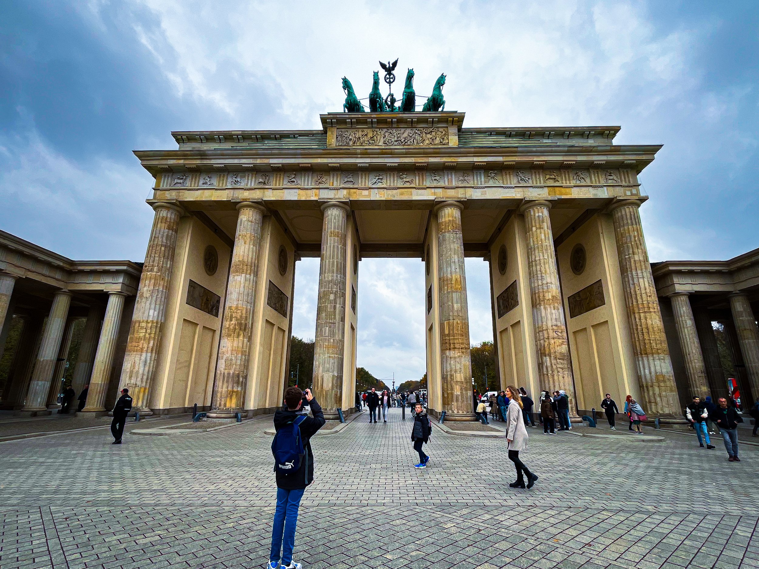The Brandenburg Gate, a historic monument in Berlin, Germany, with visitors walking around and taking pictures beneath its large columns and quadriga statue on top, visible against a cloudy sky.