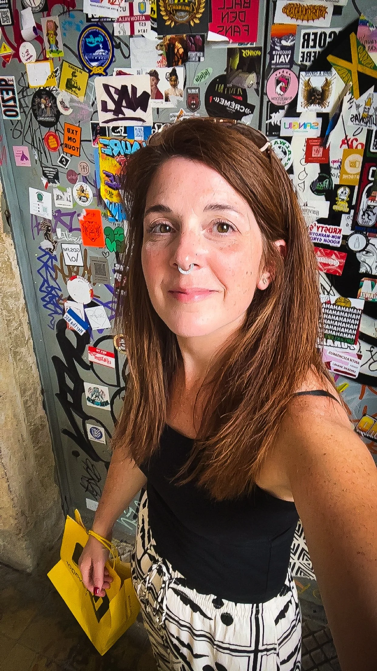 A woman with long brown hair, wearing a black tank top and black-and-white patterned skirt, taking a selfie in front of a wall covered with colorful stickers and posters. She is holding a yellow shopping bag.