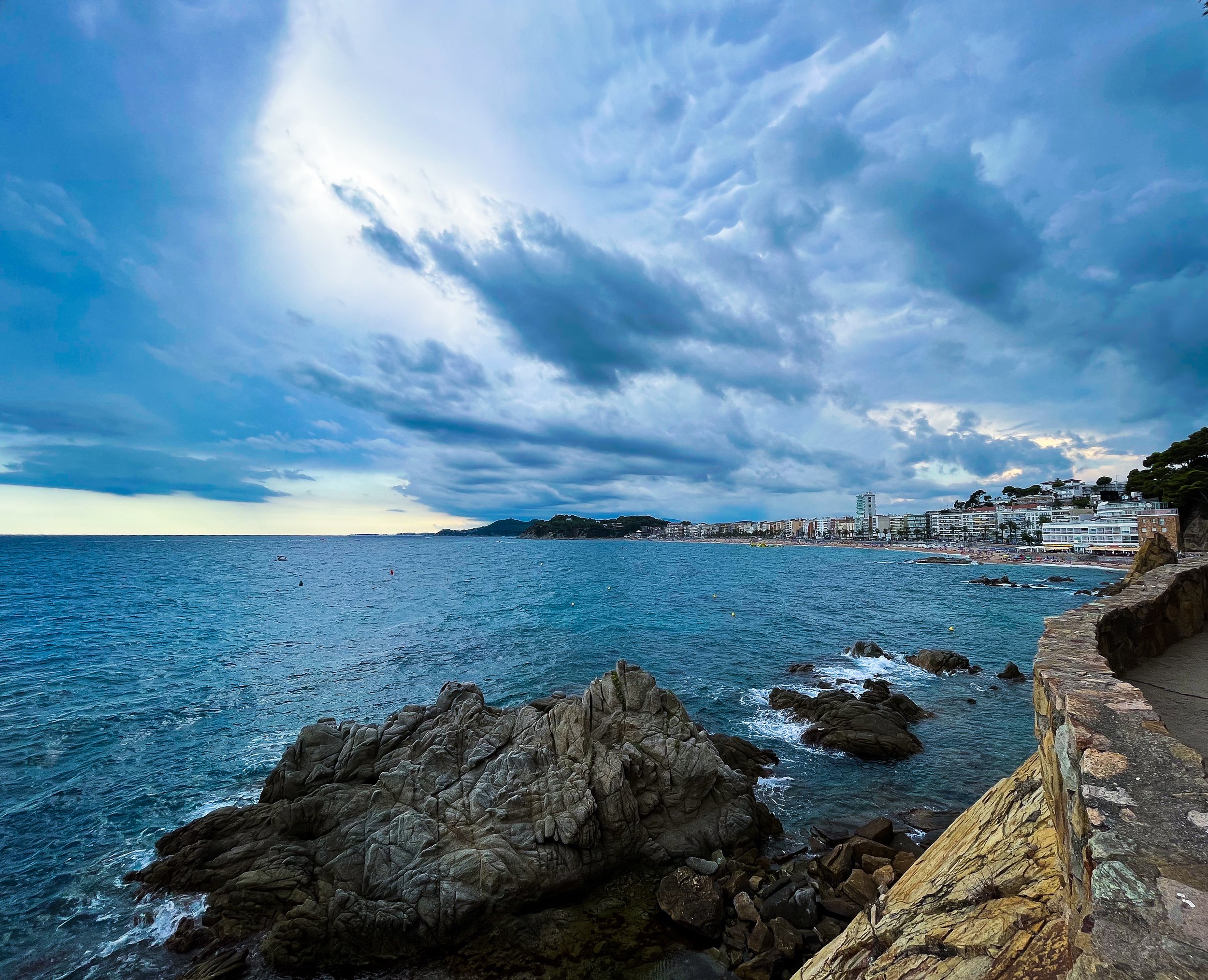 A coastal cityscape with high-rise buildings along the shoreline, under a sky filled with dark clouds, and rocky cliffs in the foreground.