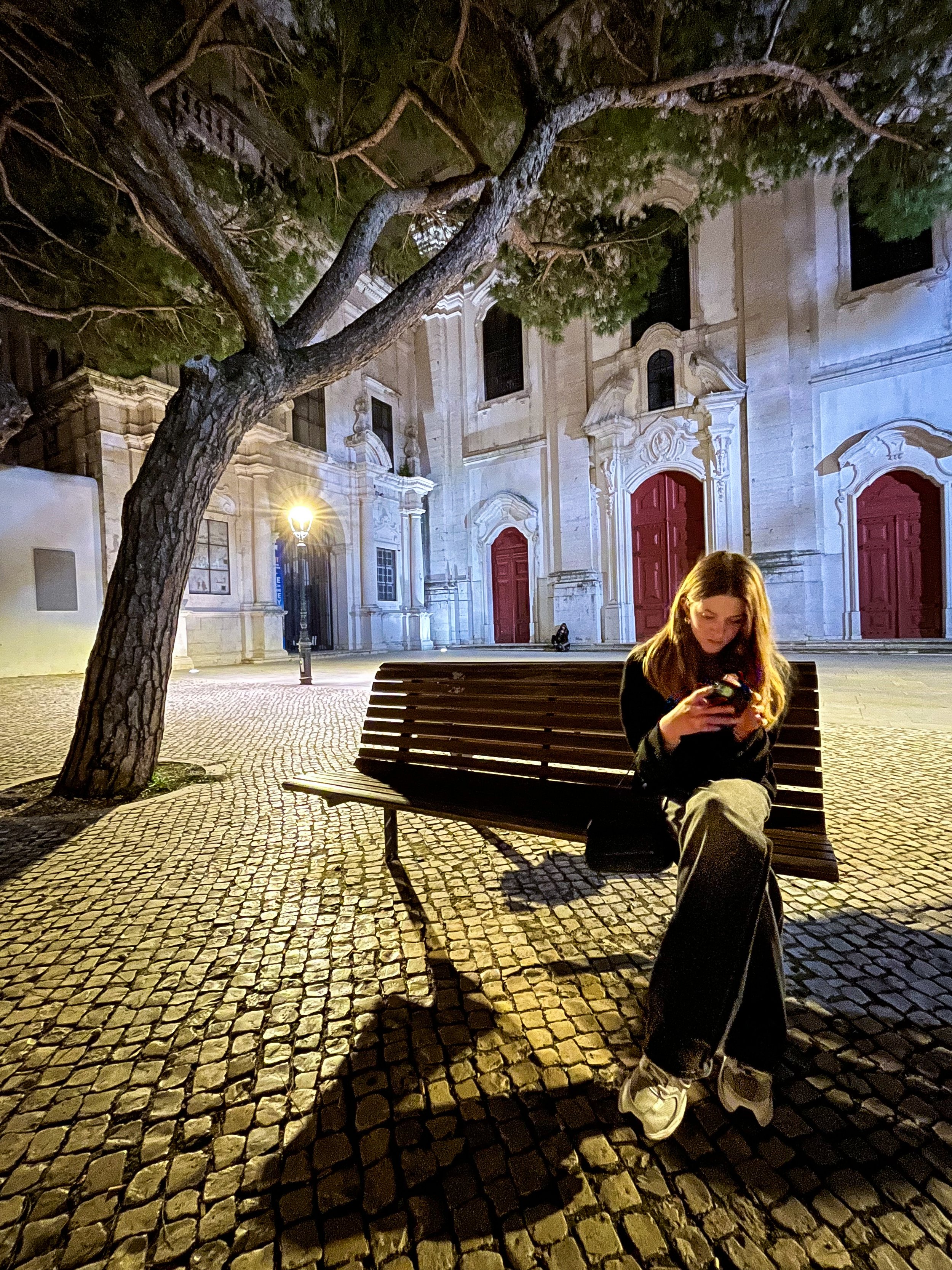 A woman with long hair sitting on a wooden bench in a historic town square at night, looking at her phone. She is wearing a black coat, camouflage pants, and white sneakers. There are cobblestone streets, a large tree, a church with ornate architecture and red doors, and some street lamps illuminating the scene.