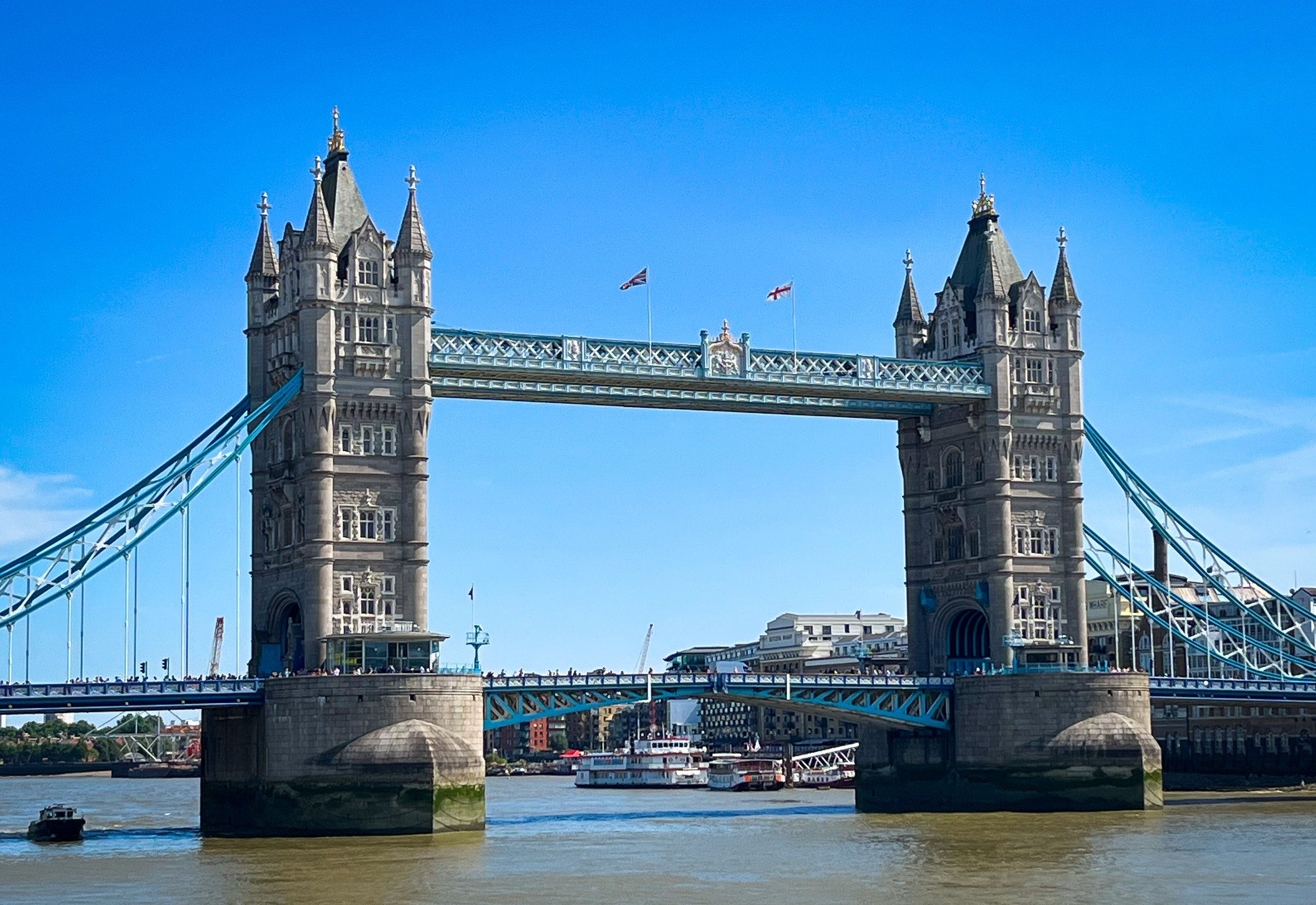 Photo of Tower Bridge in London with blue sky, flags, boats on River Thames, and buildings in background.