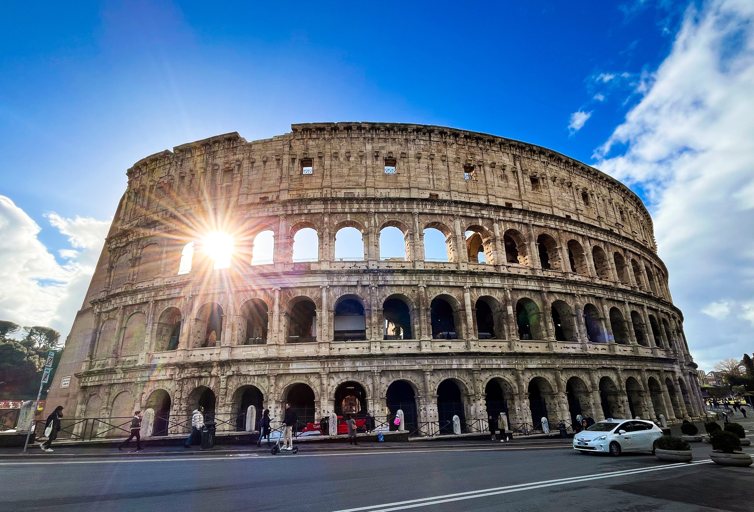 The Colosseum in Rome, Italy, with the sun shining behind it, blue sky with some clouds, and people walking on the street in front.