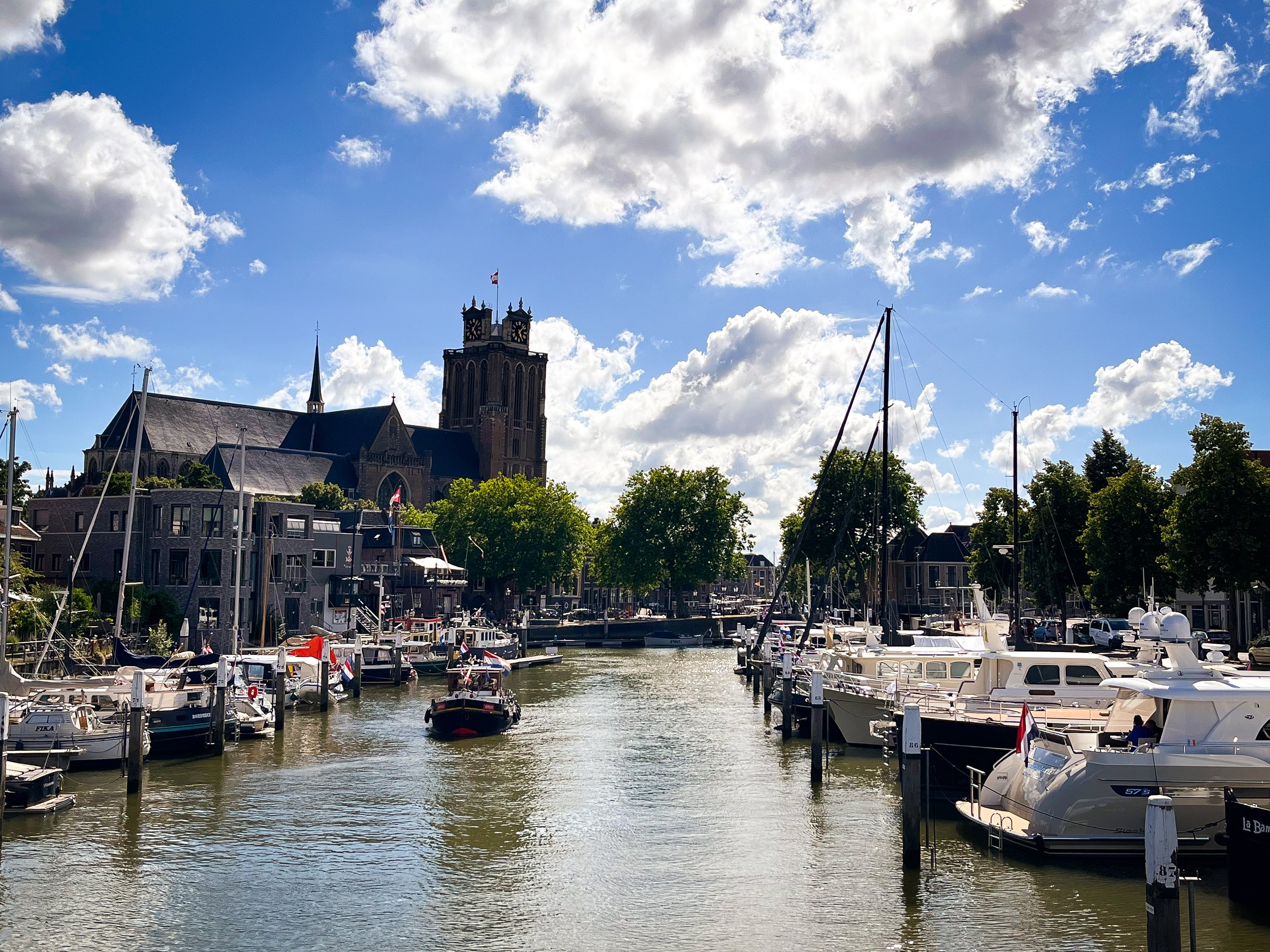 A marina with boats docked on the water, a church or historic building with a clock tower in the background, and partly cloudy skies.