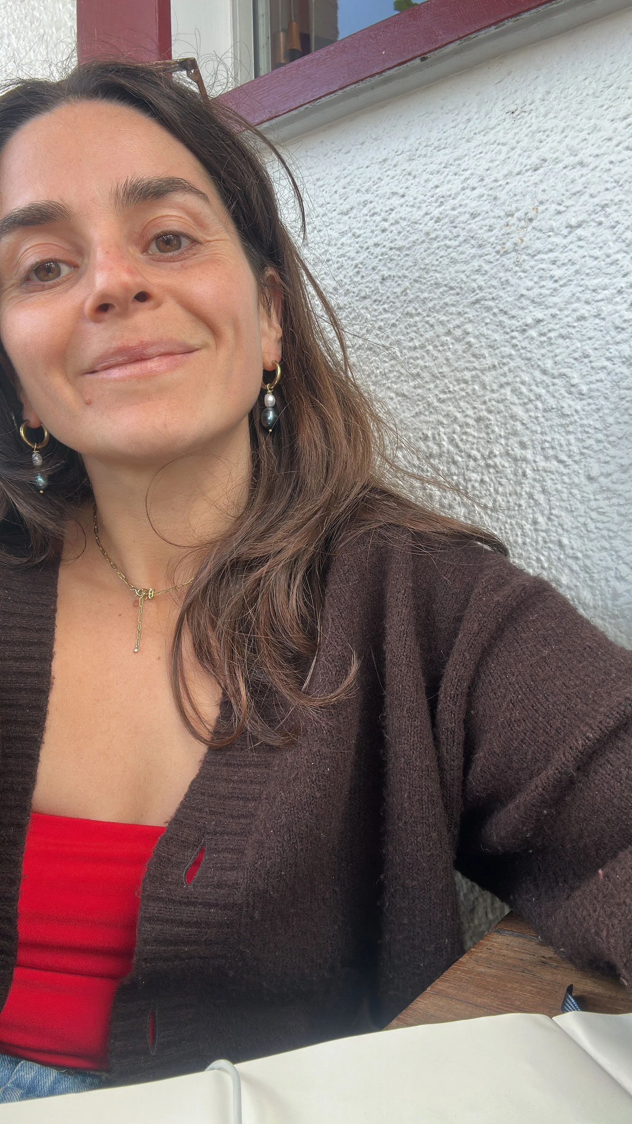 A woman with brown hair, wearing earrings with pearls, a necklace, a red top, and a brown cardigan, sitting outside near a white wall with a window and a table.