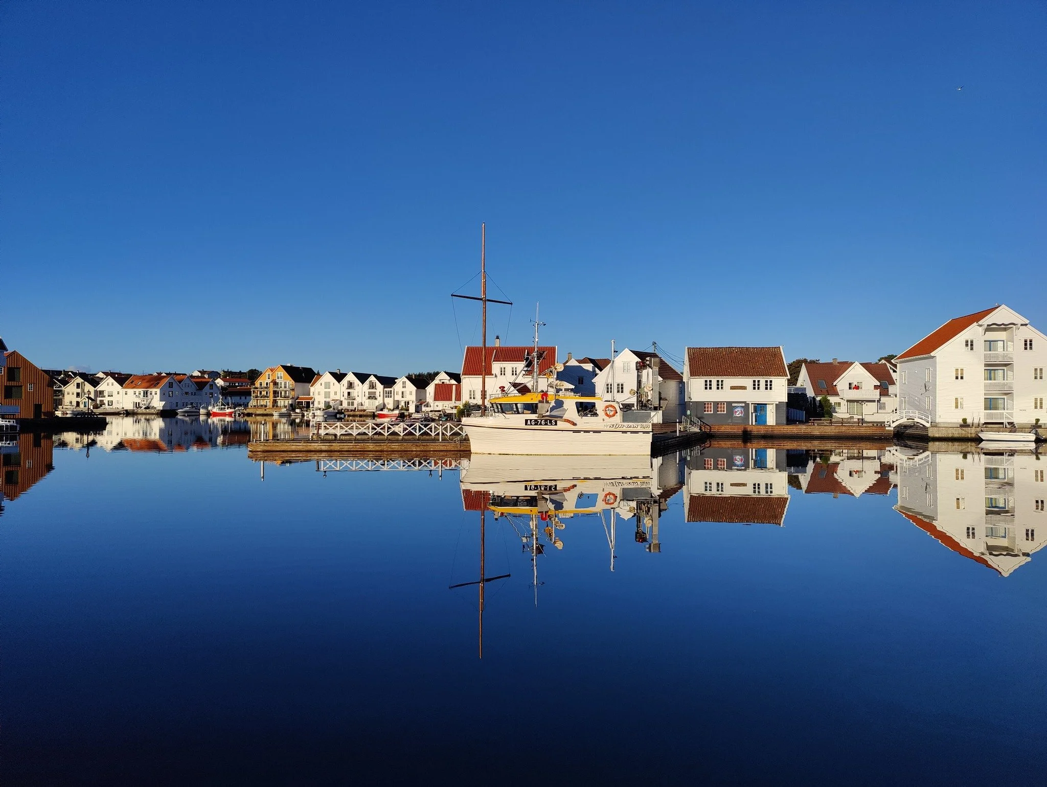 Byskjermet havn med fartøy og hus, reflektert i vannet under en klar blå himmel.