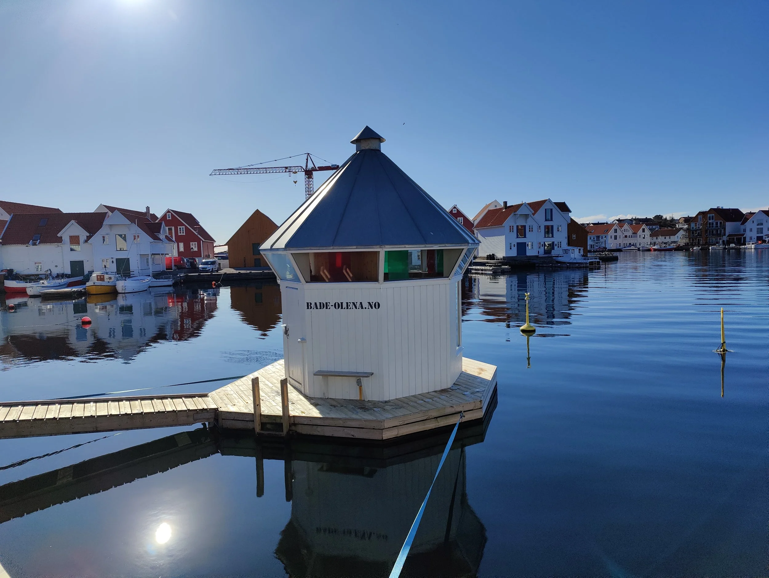 En hvit liten bygning med blå tak som ligger på en trebrygge i en stille havn med flere hus i bakgrunnen, og reflektert i vannet.