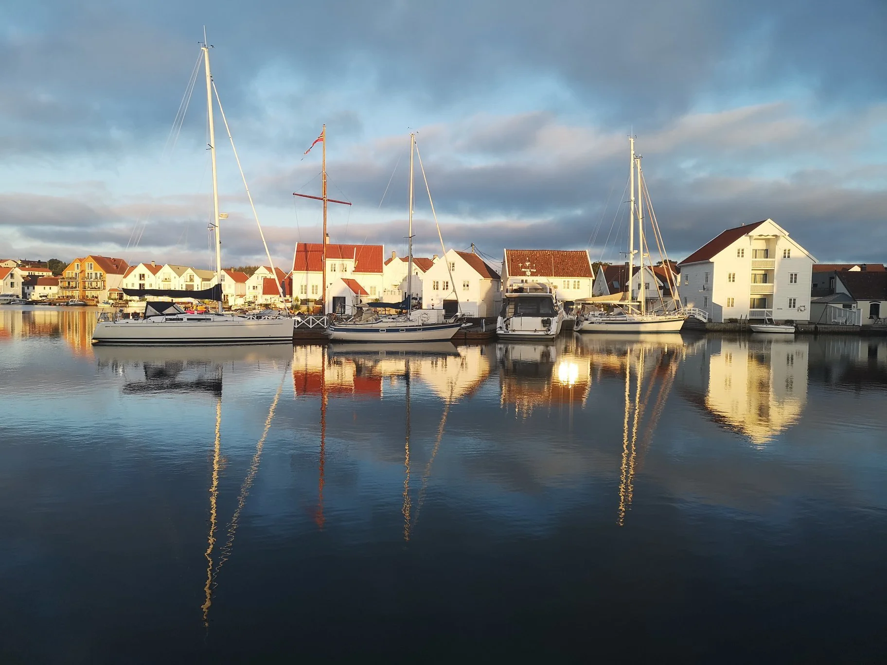 Båter ved brygga i et rolig vann, med hus i bakgrunnen og overskyet himmel.