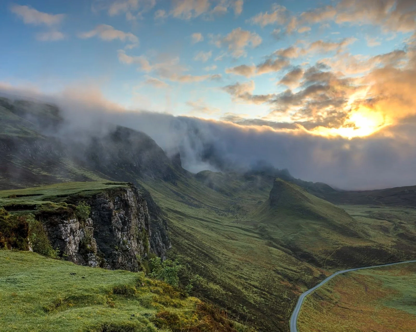 A scenic landscape of green rolling hills and rocky cliffs with a misty mountain waterfall in the background and a winding road in the foreground, during sunset with clouds and sunlight.