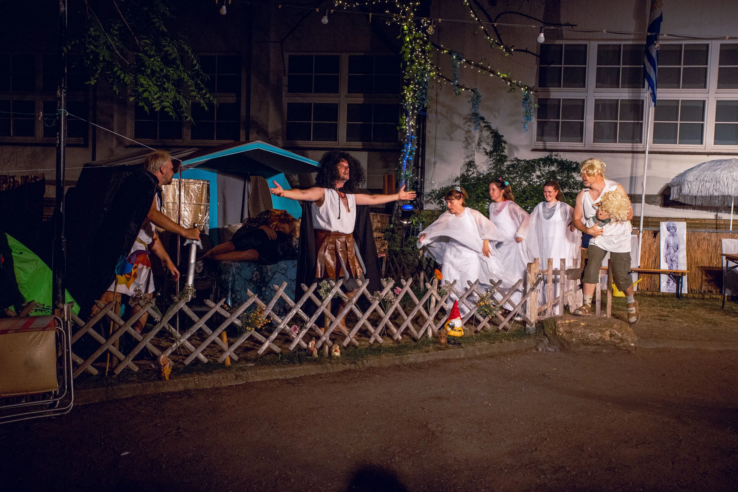 Theateraufführung einer Gruppe von Menschen in Kostümen bei Nacht, im Freien, vor einem improvisierten Bühnenbild mit Lichterketten und Dekorationen.