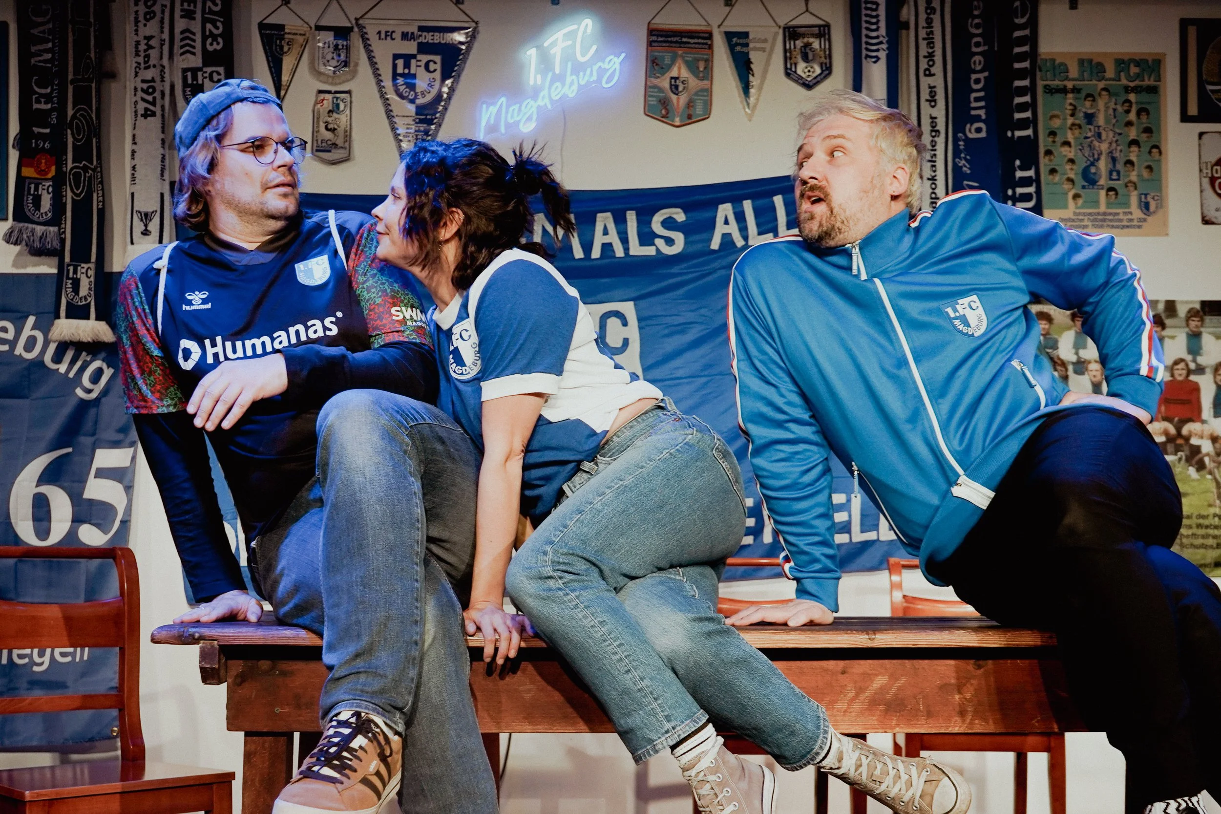 Drei Personen in einem Raum, der mit Fußball- und Fanartikeln dekoriert ist, sitzen auf einem Holztisch und scheinen in einem Gespräch vertieft zu sein. Sie tragen sportliche Kleidung mit dem Logo des 1. FC Magdeburg.