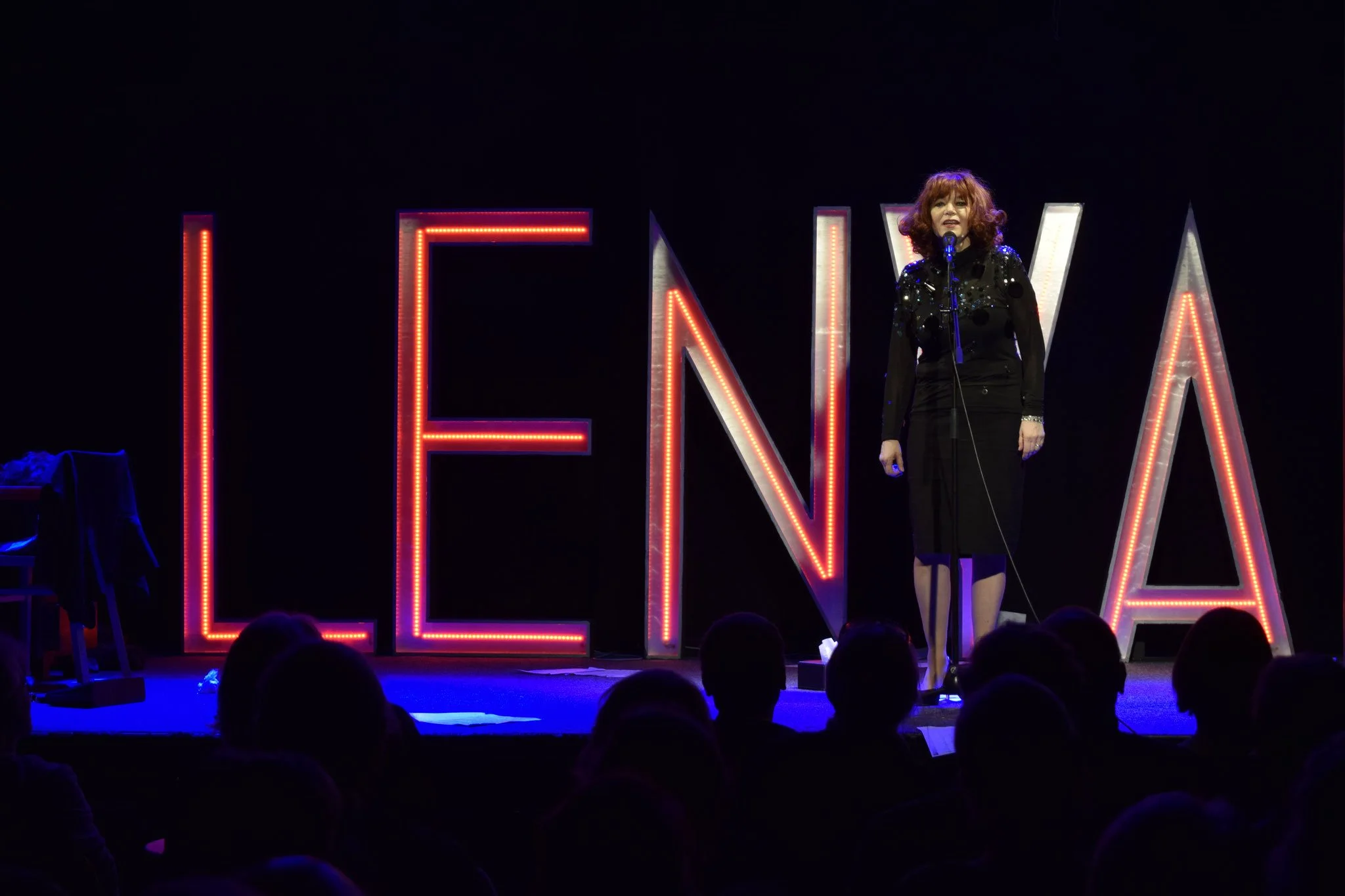 Eine Frau steht mit einem Mikrofon auf einer Bühne vor großen, leuchtenden Buchstaben, die L, E, N, A bilden. Die Bühne ist dunkel, und das Publikum schaut zu.