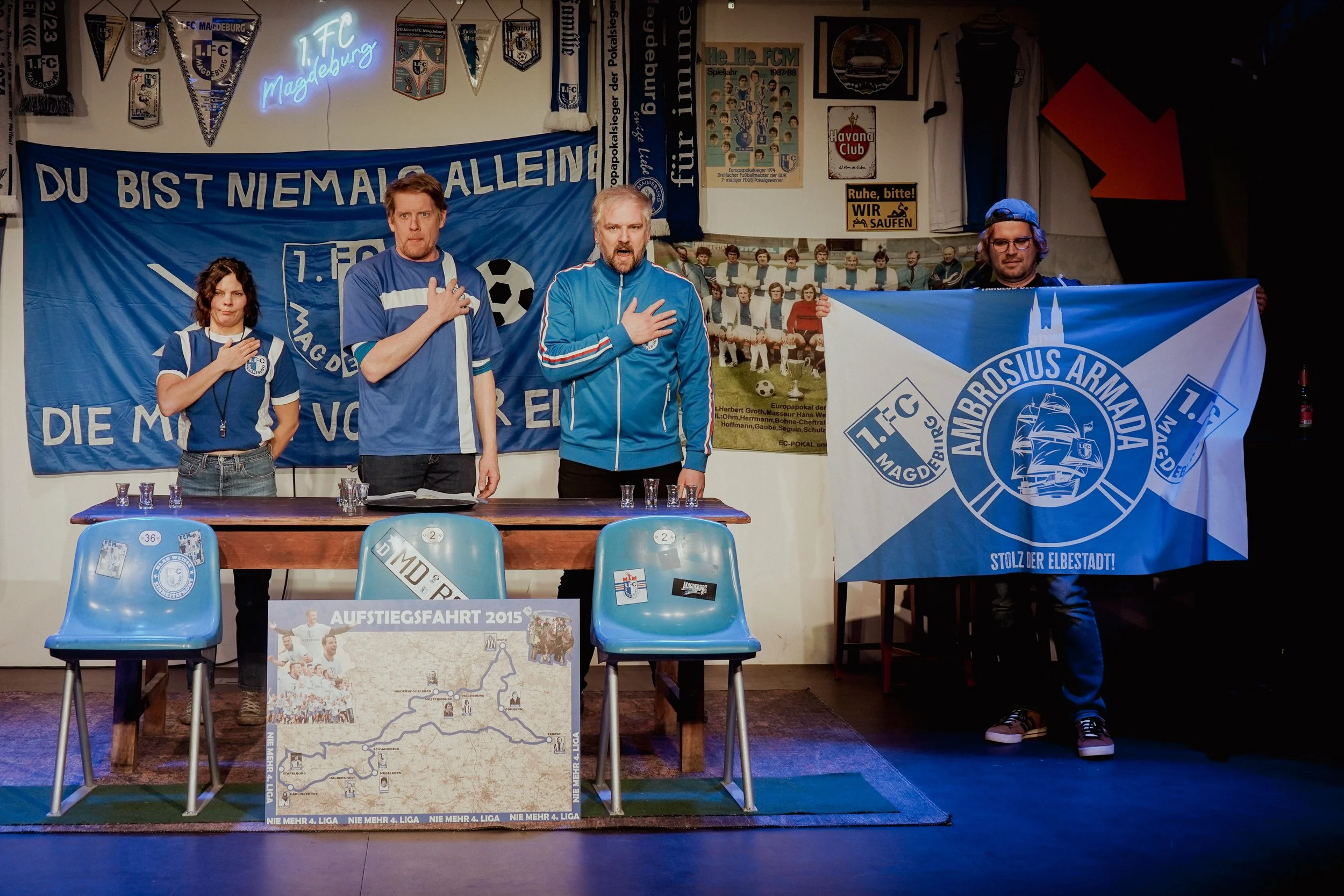 Drei Personen in blauen Sportjacken stehen an einem Tisch, halten die rechte Hand auf die Brust und machen den Hitlergruß, während eine Person rechts einen blauen und weißen FC-Magdeburg-Flagge hält. Im Hintergrund sind blaue Fahnen, Poster und Fotos
