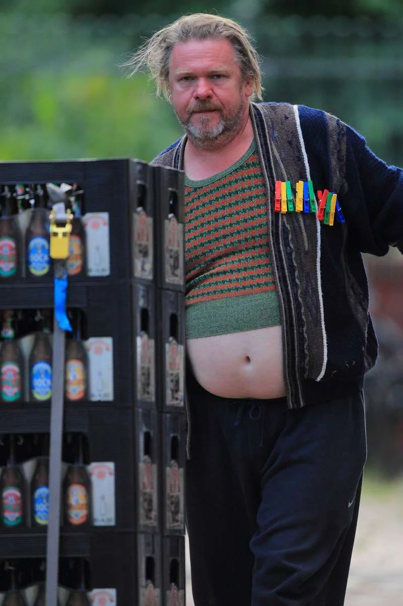 Ein Mann mit zerzaustem Haar und Bart, trägt eine bunte Jacke und ein grünes Hemd, das darüber hervorscheint, steht neben einem Getränkekasten, der mehrere Bierflaschen enthält.