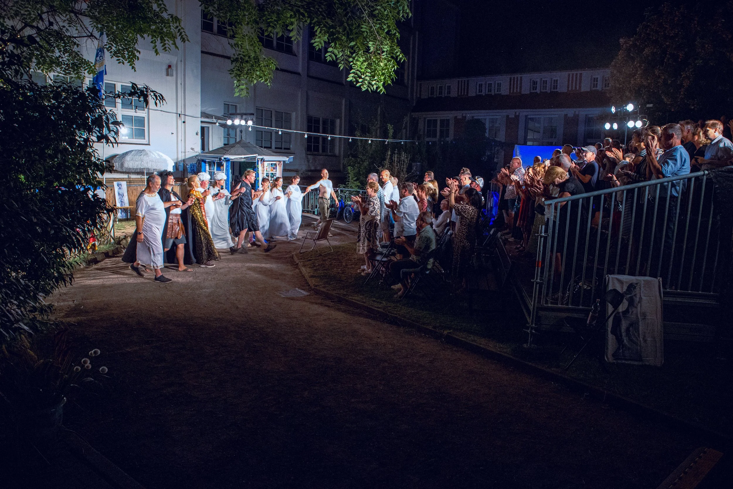 Eine Gruppe von Menschen tanzt draußen bei Nacht, während eine weitere Gruppe auf einer Tribüne zuschaut. Es gibt Lichter, Bäume und Gebäude im Hintergrund.
