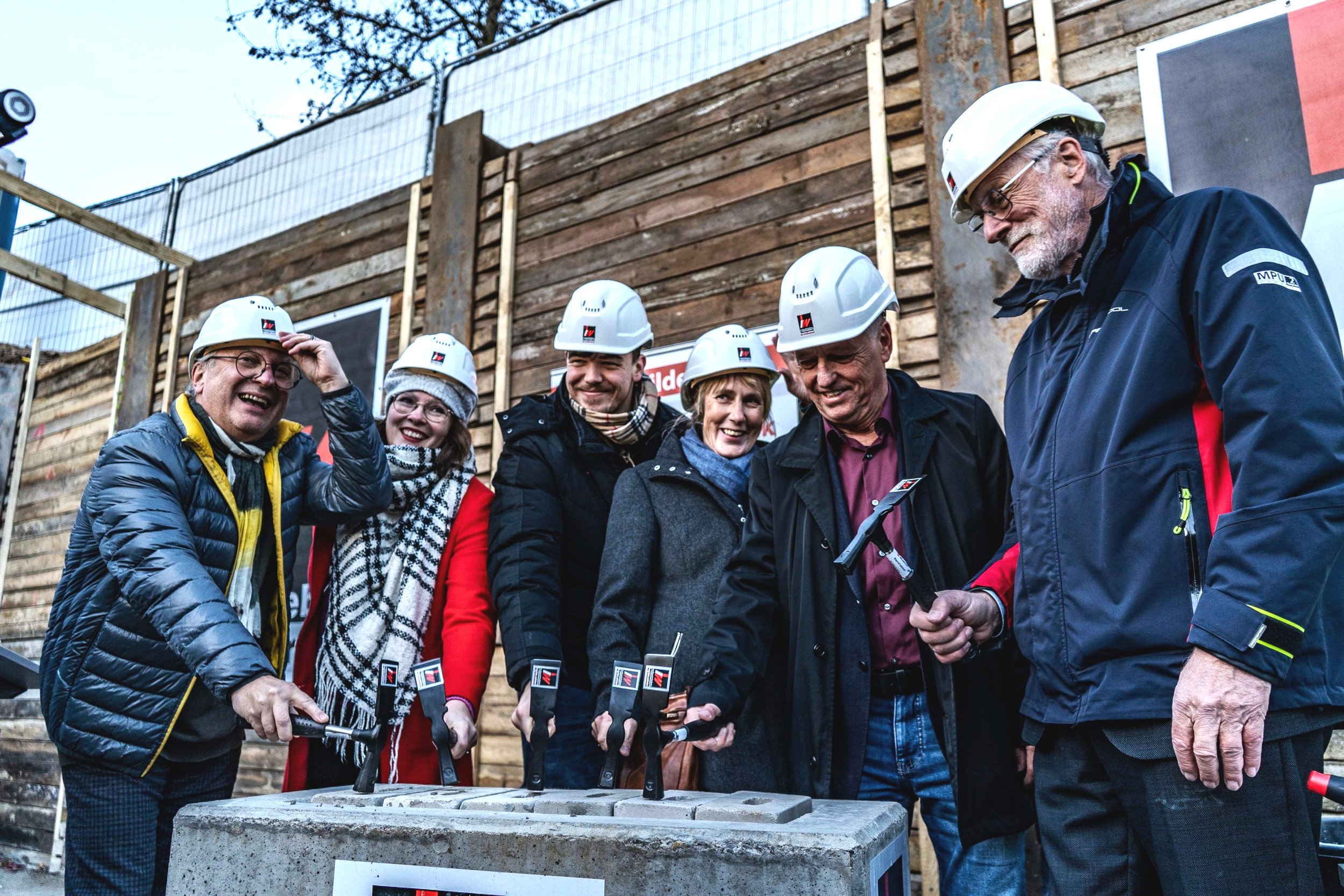 Gruppen von Feiernden mit Schutzhelmen bei einer Baubegehung, die einen Betonkern durch Hämmern enthüllen.