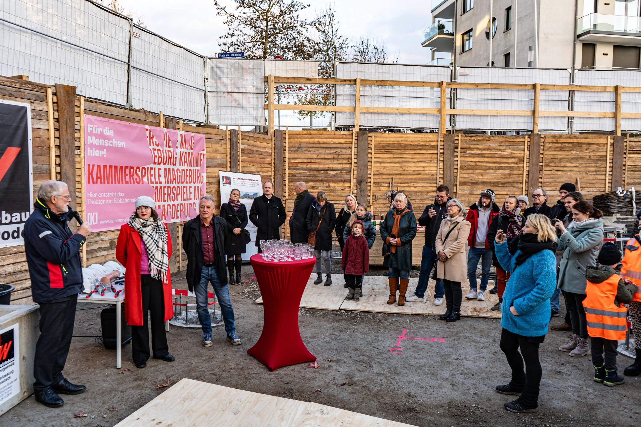 Eine Gruppe von Menschen steht auf einer Baustelle, während ein Mann mit einem Mikrofon spricht. Es gibt eine große pinkfarbene Werbetafel im Hintergrund. Einige Menschen filmen oder fotografieren die Szene, andere hören zu.
