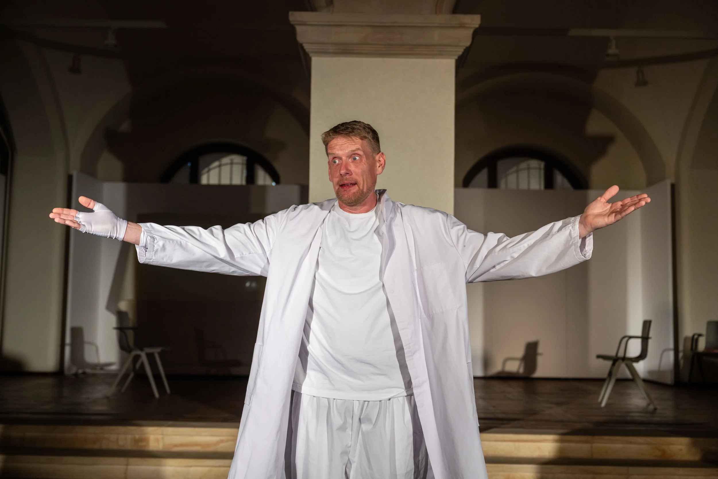 Einmann-Theater mit Person in weißem Arztkittel, der breite Armlage macht, vor einem Bühnenraum