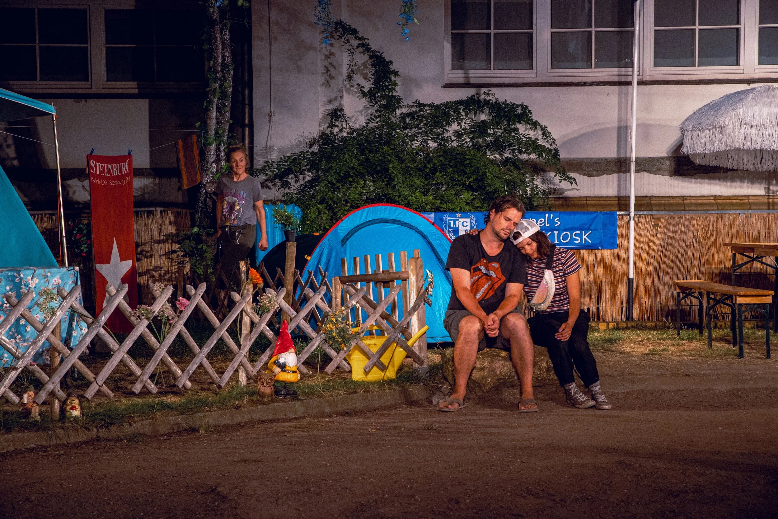 Zwei Personen sitzen am Abend vor einem Zelt auf einem Campingplatz, während eine Frau im Hintergrund hinter einem Gartenzaun steht. Das Zelt ist blau, umgeben von Blumen und Dekoartikeln.