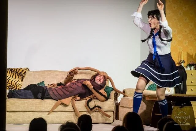 Auf einer Bühne 2 Frauen, eine liegt auf einem Sofa, die andere steht und schreit. Die Frau auf dem Sofa hält eine große, künstliche Spinne. Die stehende Frau trägt ein Schuluniform-Outfit.
