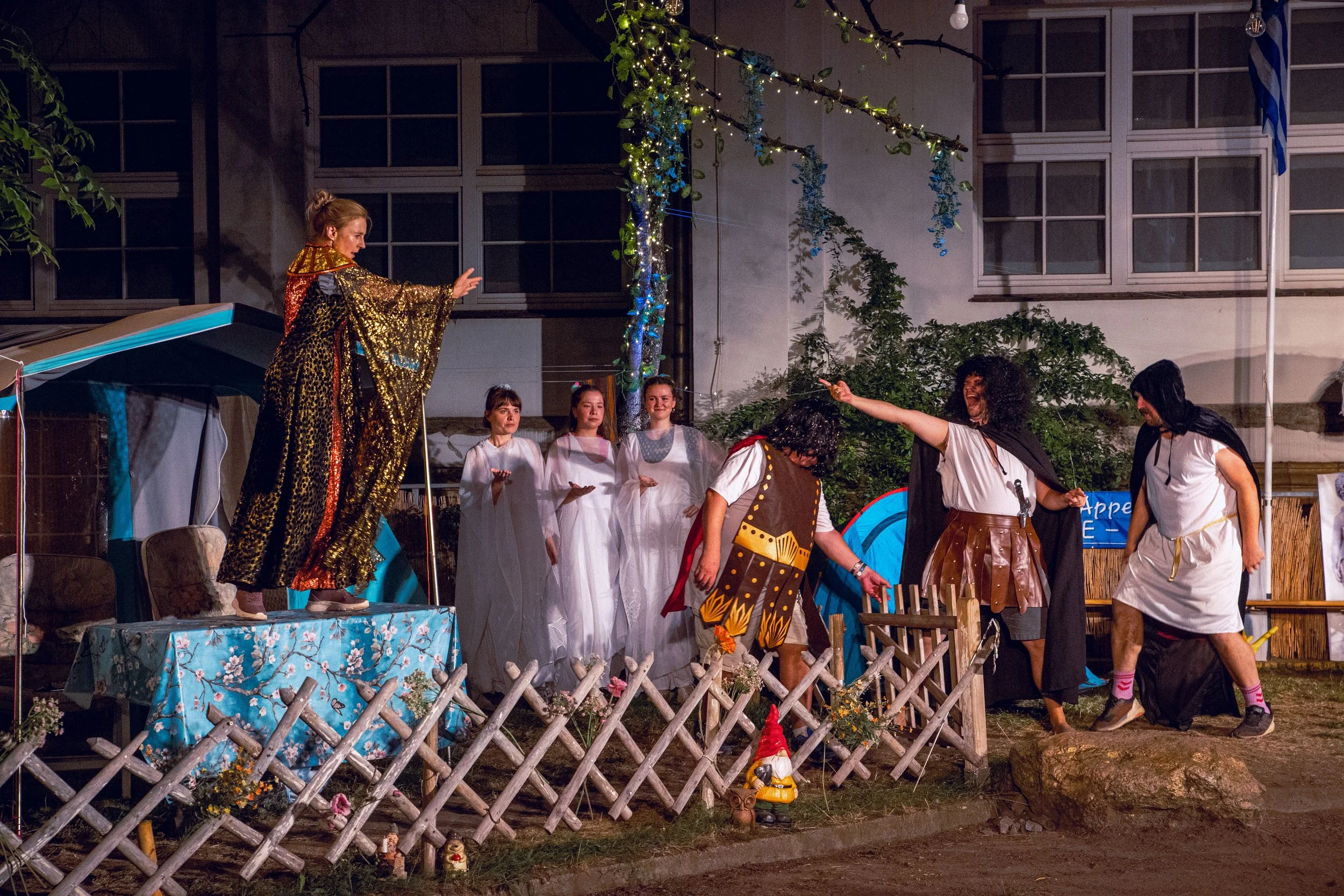 Szenen aus einem Theaterstück, bei dem Menschen in Kostümen eine Szene auf einer kleinen Bühne im Freien darstellen. Eine Person in goldfarbenem Umhang steht erhöht, während andere in weiße und bunte Kostüme eingebunden sind.