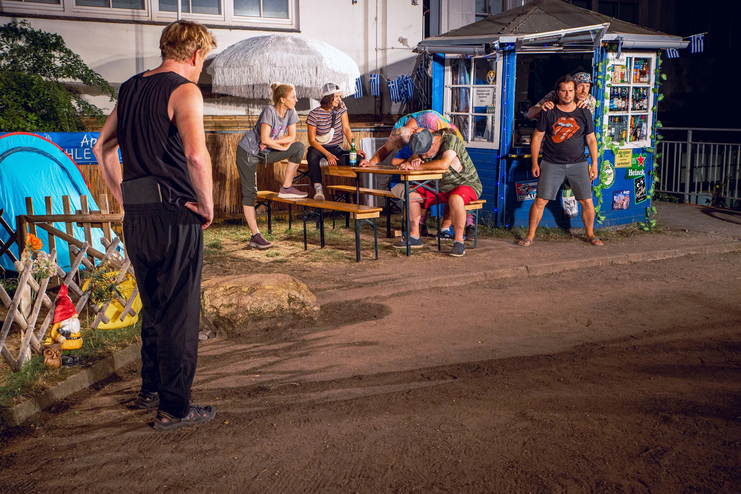 Menschen sitzen und stehen vor einem kleinen blaue Verkaufsstand bei Nacht, einige sind erschöpft, eine Person schläft auf einem Tisch, während andere sich unterhalten oder die Umgebung beobachten.