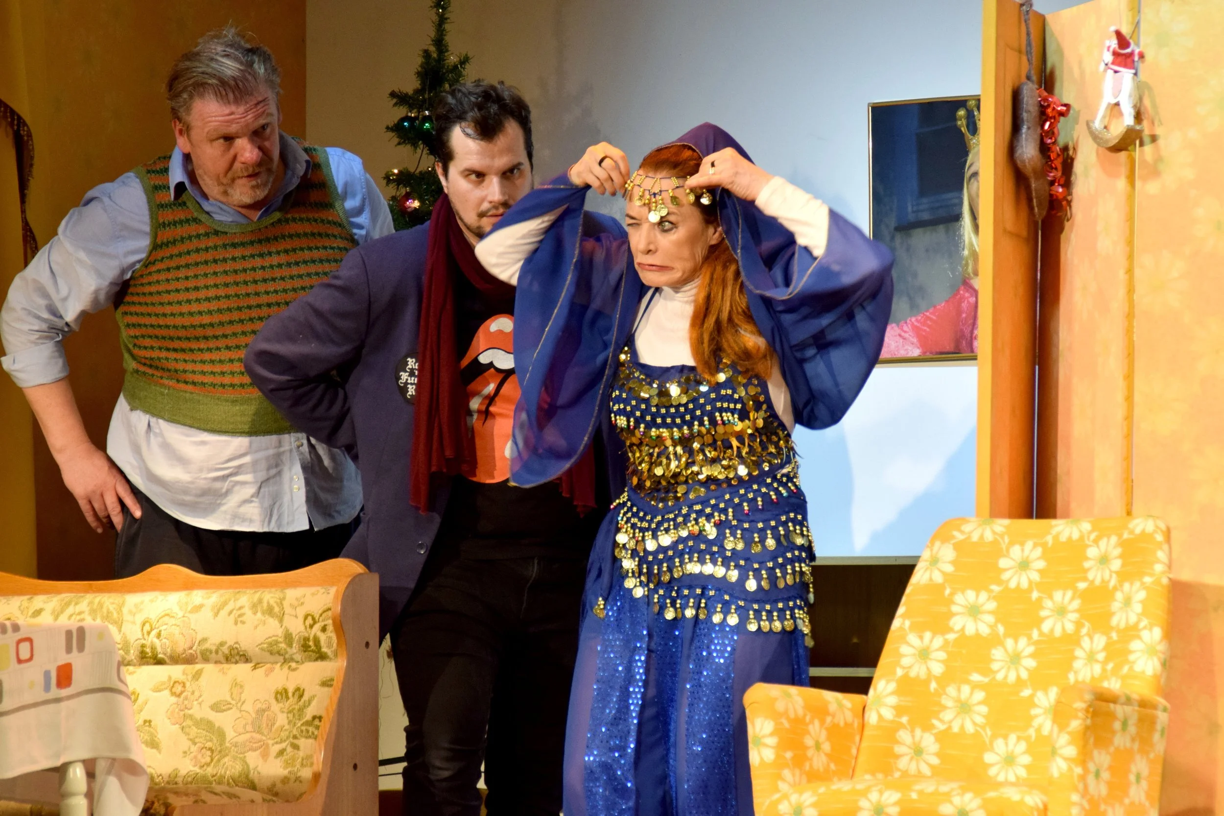 Drei Männer und eine Frau in einem Wohnzimmer, die eine Tanz- oder Theaterperformance machen, mit Weihnachtsdekoration im Hintergrund, die Frau trägt ein blaues, glitzerndes Kostüm und eine goldene Kopfbedeckung.