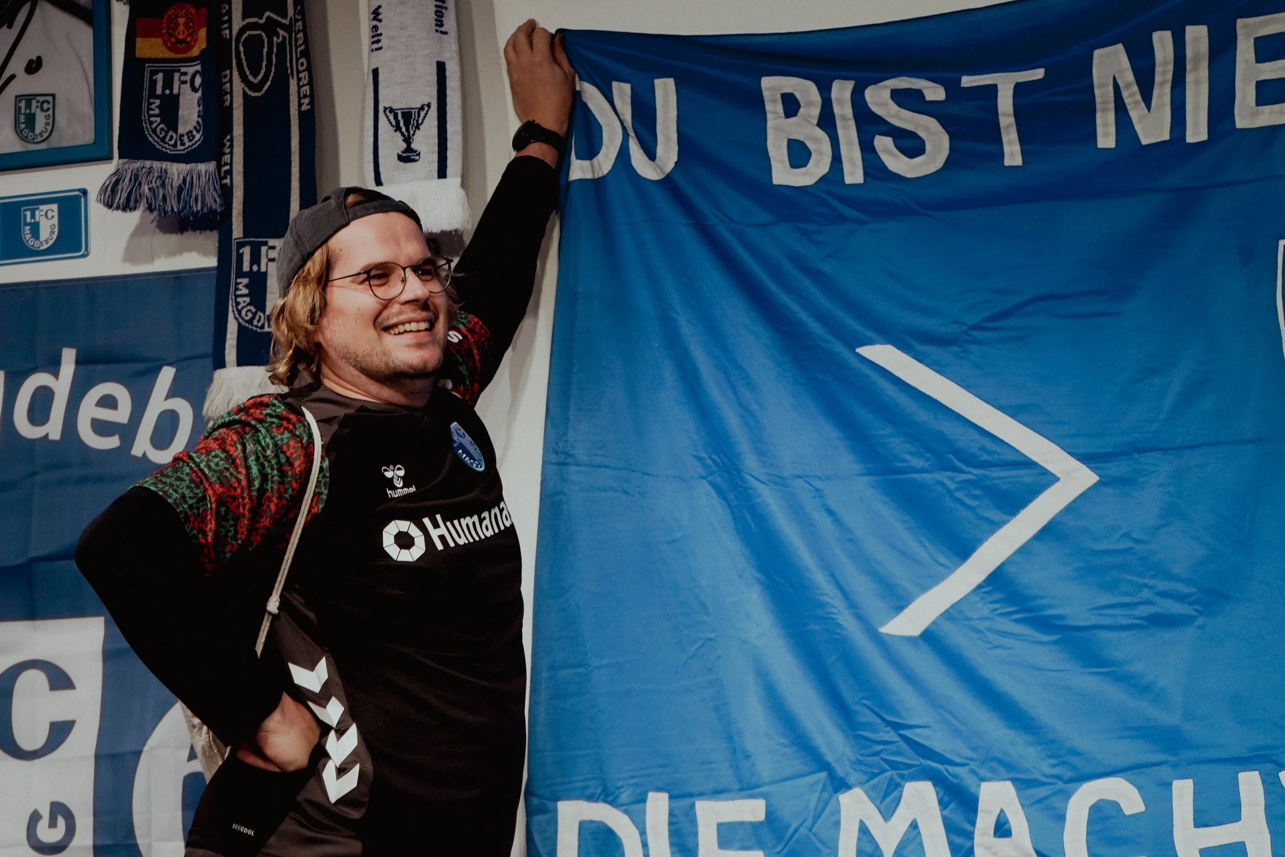 Ein Mann mit Brille, schwarzem Cap und einem schwarzen Trikot, lächelnd, steht vor einer blauen Fahne mit einem weißen Pfeilsymbol, umgeben von weiteren Fahnen und Schals, die mit Sportvereinen und -logos geschmückt sind.