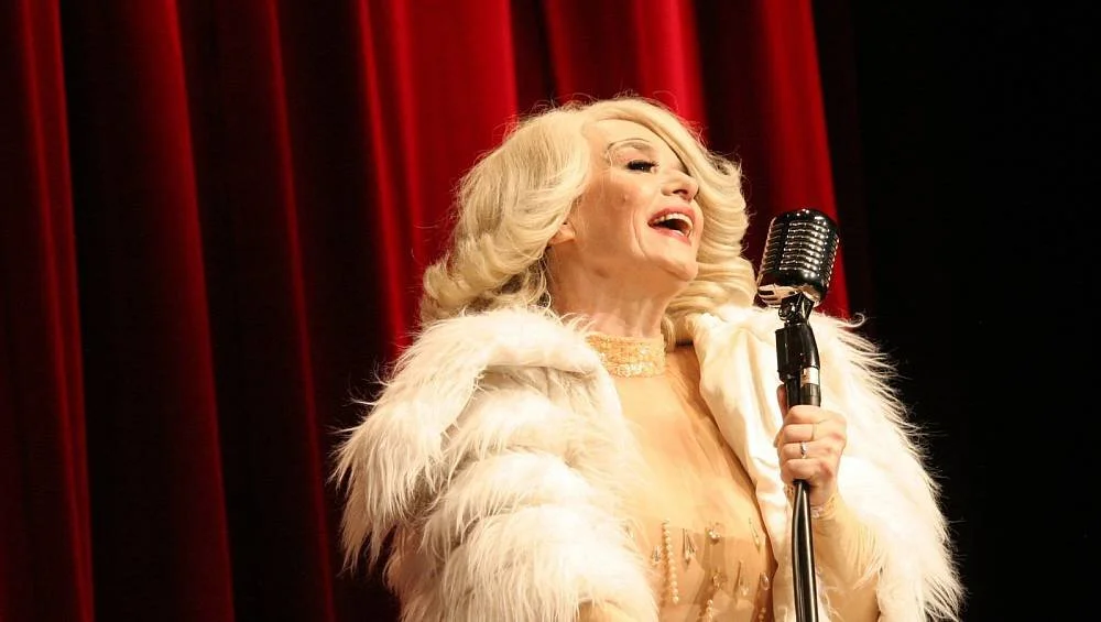 Eine ältere Frau mit blonden, lockigen Haaren singt vor rot-gestreiften Vorhängen und hält ein Mikrofon in der Hand.
