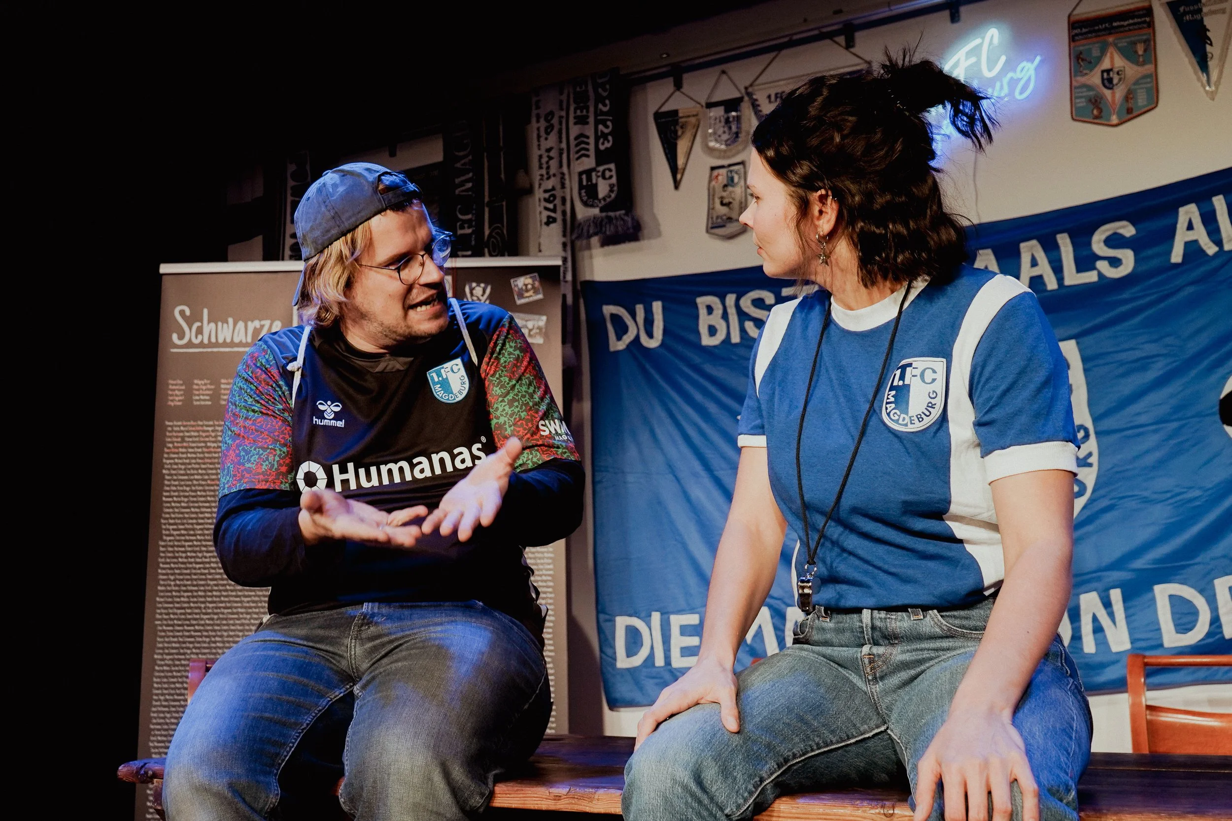 Zwei Personen sitzen auf einer Bank im Gespräch, beide tragen blaue Trikots mit einem Fußballverein-Logo, im Hintergrund Vereinsbanner und Poster.
