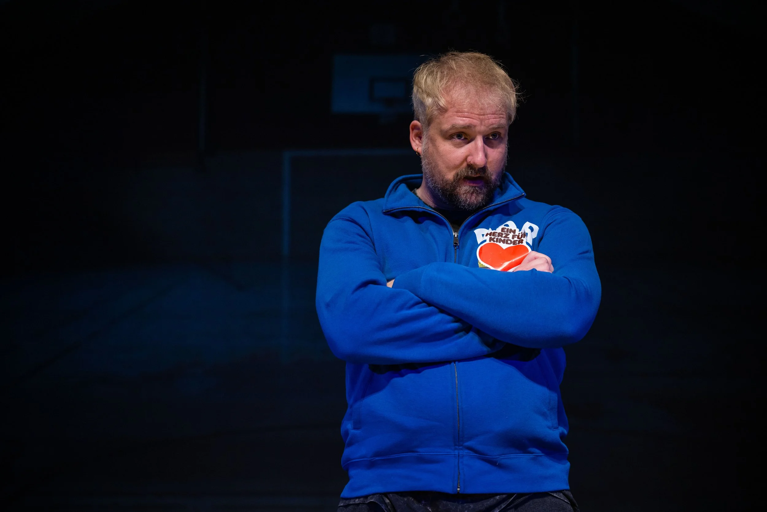 Mann in blauer Jacke mit verschränkten Armen auf einer dunklen Bühne