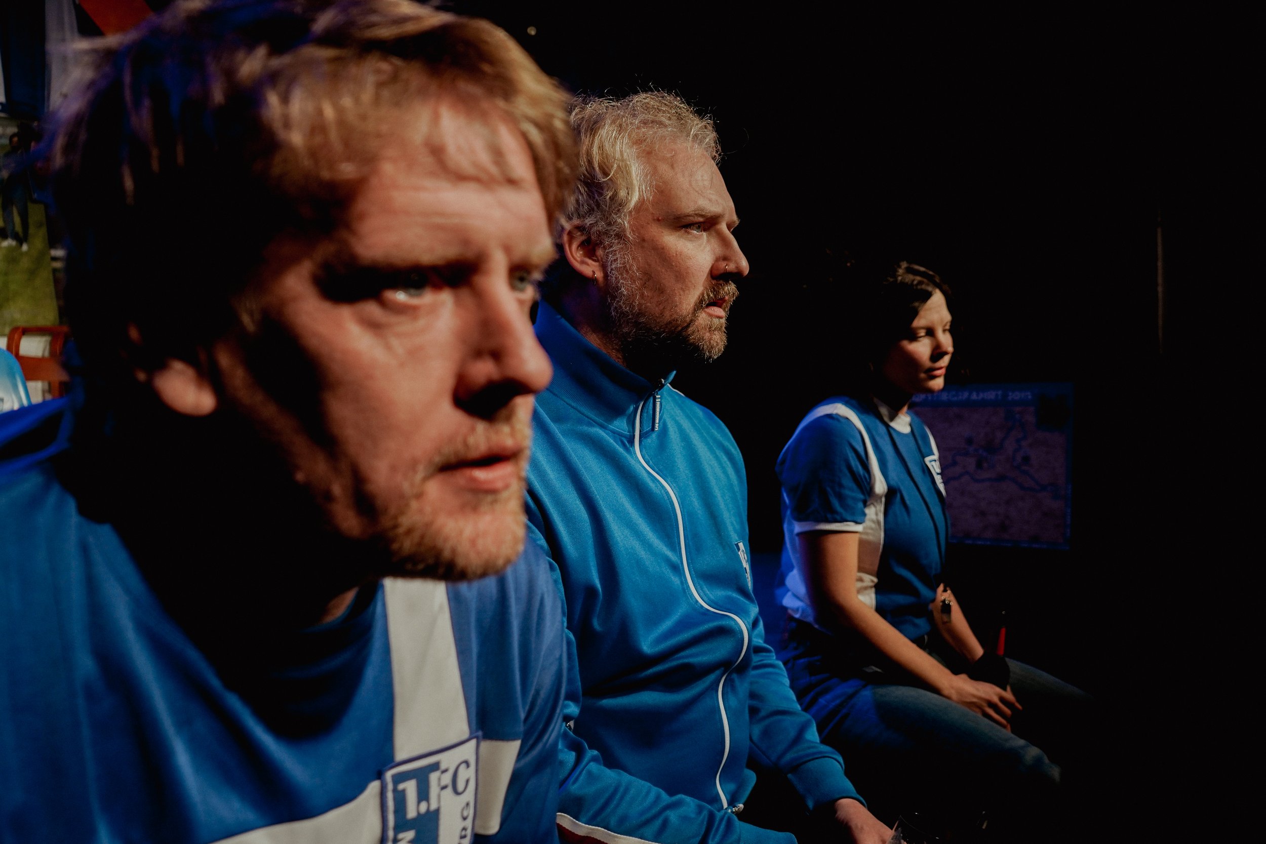 Drei Menschen in blauen Sportjacken sitzen nebeneinander im Dunkeln, im Hintergrund ist ein Bildschirm zu erkennen.