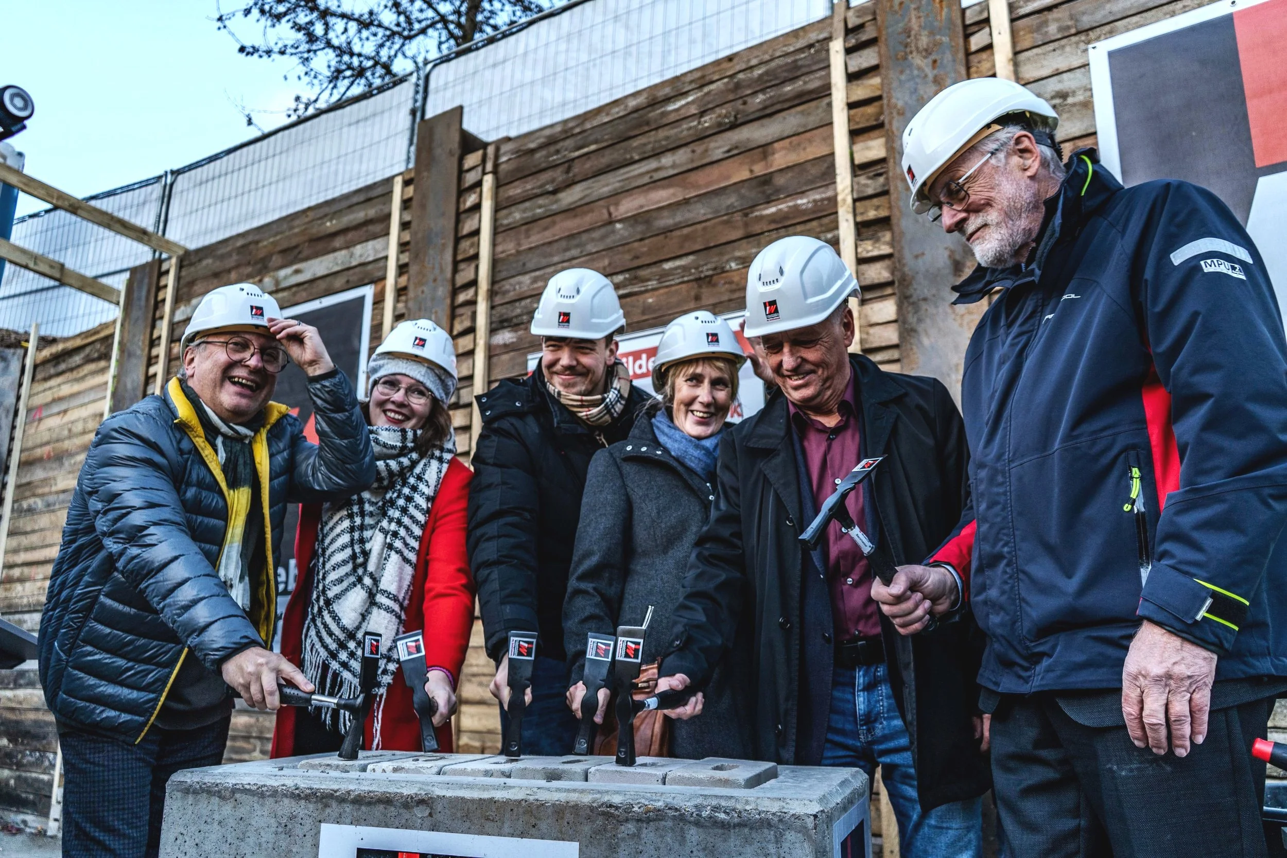 Gruppe von sechs Menschen bei einer Bauarbeitszeremonie, alle tragen Helme, Steine und Werkzeuge in der Hand, lächelnd vor einem Holzgerüst.