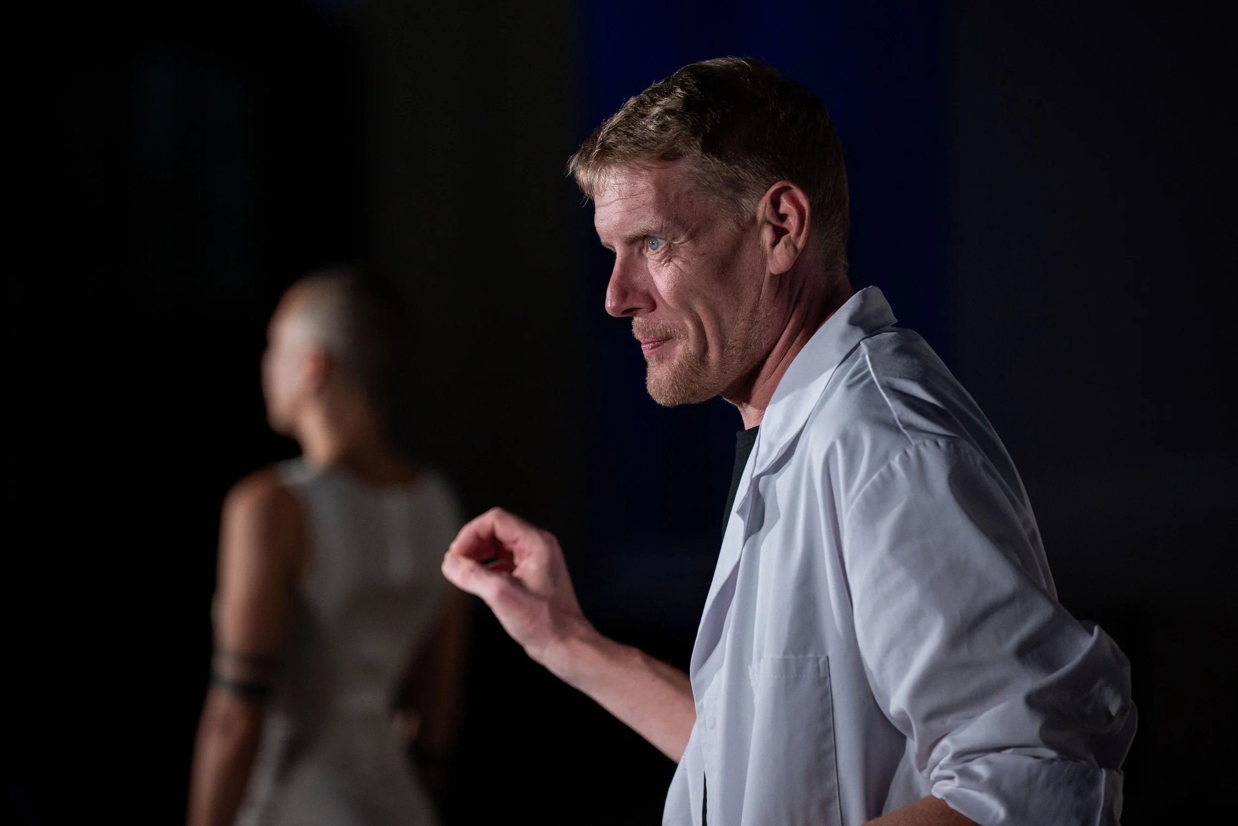 Ein Mann in weißem Arztkittel befindet sich im Profil auf der rechten Seite des Bildes, im Hintergrund ist eine Frau in einem weißen Kleid und mit gefärbtem Arm zu sehen, sie ist unscharf.