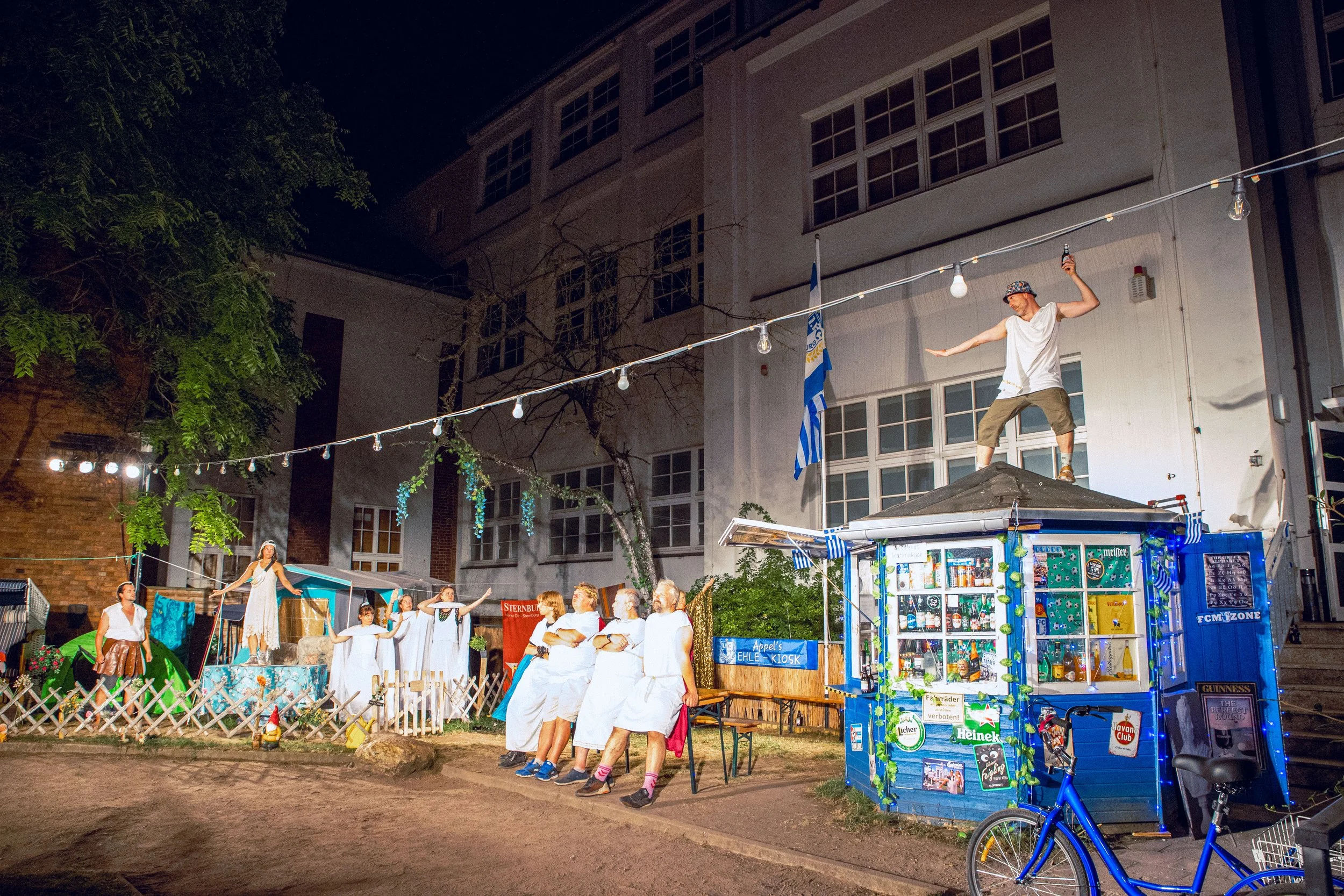 Bodenständiges Theaterstück mit Menschen in weißen Kleidern, die auf einer Bühne in einem Innenhof beim Nacht aufgeführt wird, umgeben von einer kleinen Menschengruppe, einer Fassade eines Gebäudes im Hintergrund und einem blauen Kiosk mit Weihnachts