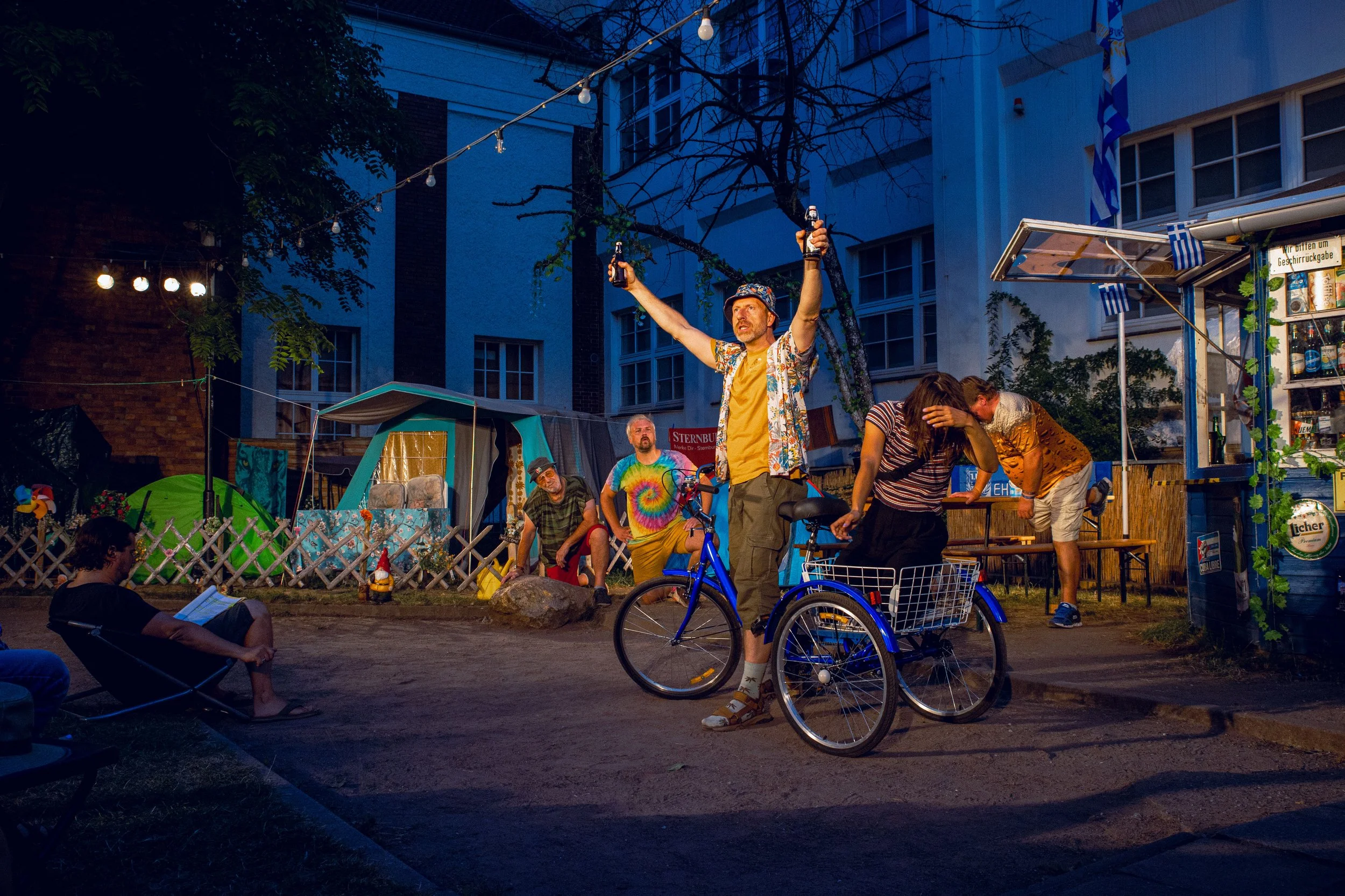 Menschen beim öffentlichen Fest am Abend, mit einem Mann in bunter Sommerhose und Fahrrad im Mittelpunkt, umgeben von weiteren Personen, Zelten und Lichterketten.