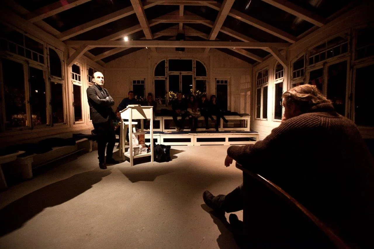 Eine Person steht an einem kleinen Podium und spricht vor einer Gruppe von Zuhörern in einem lichtdurchfluteten Holzzimmer mit großen Fenstern, sitzend in einem Kreis.