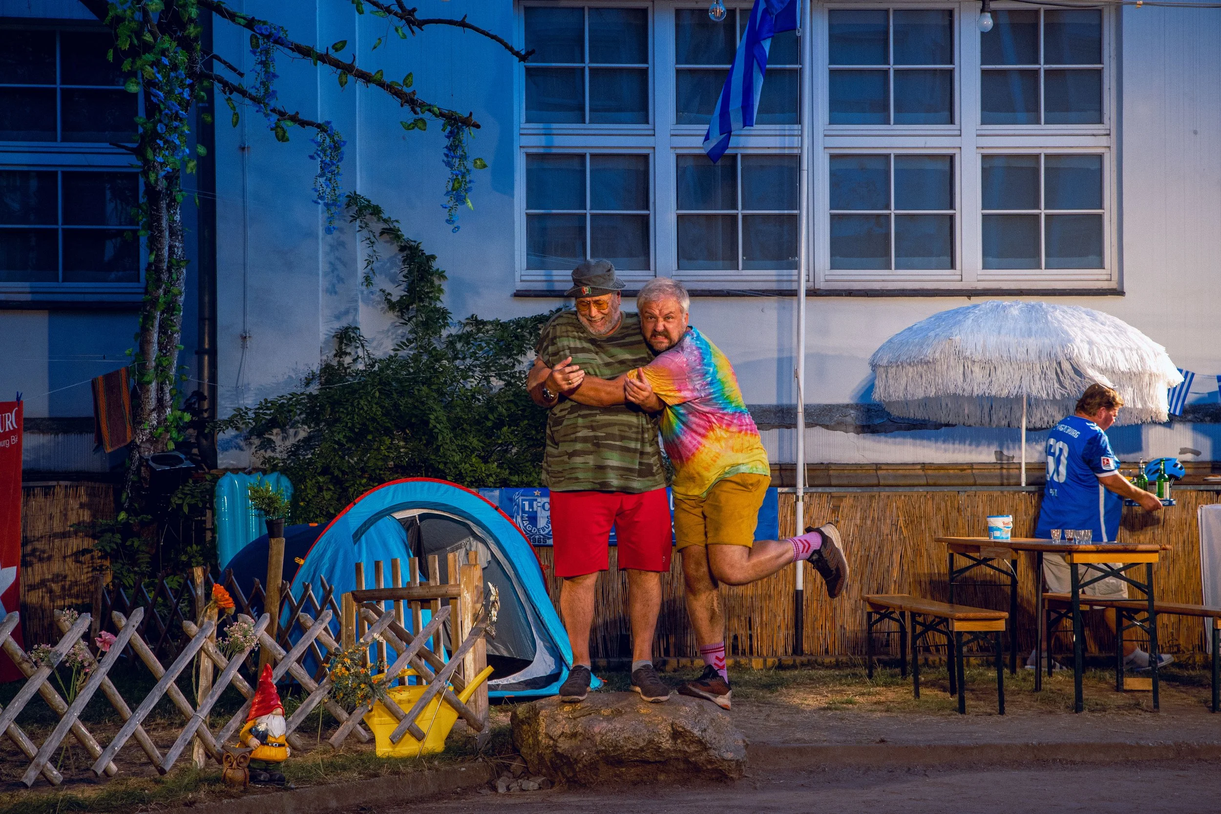 Zwei Männer, einer in Camouflage-Shirt und rotem Shorts, der andere in buntem T-Shirt und gelben Shorts, posieren lächelnd auf einem Stein. Im Hintergrund steht eine Party im Freien mit Zelt, Sonnenschirm, Getränke und anderen Personen.